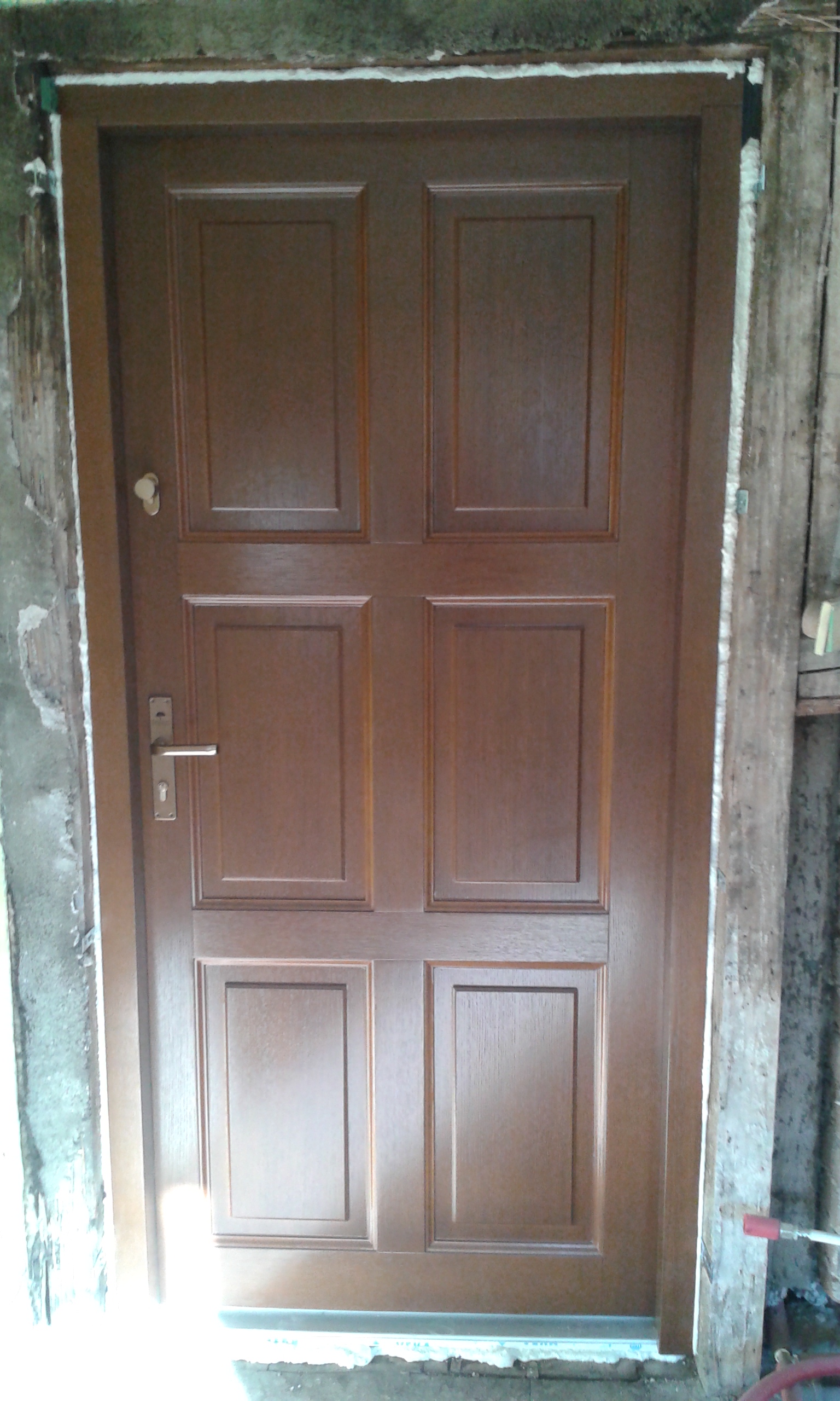 Drzwi wejściowe osadzone w otworze z widocznymi warstwami montażowymi i nieukończonym ościeżem, oczekujące na finalne prace wykończeniowe.