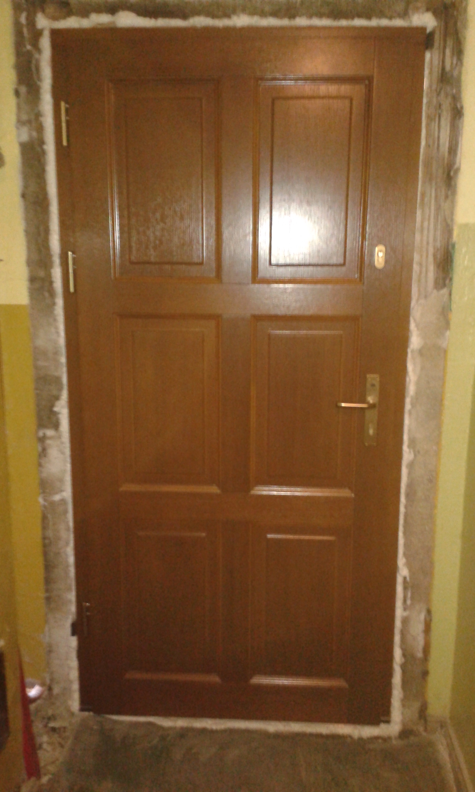 Drzwi wejściowe osadzone w nieukończonym otworze, z widocznymi śladami prac montażowych i przygotowaniem ościeża do dalszego wykończenia.