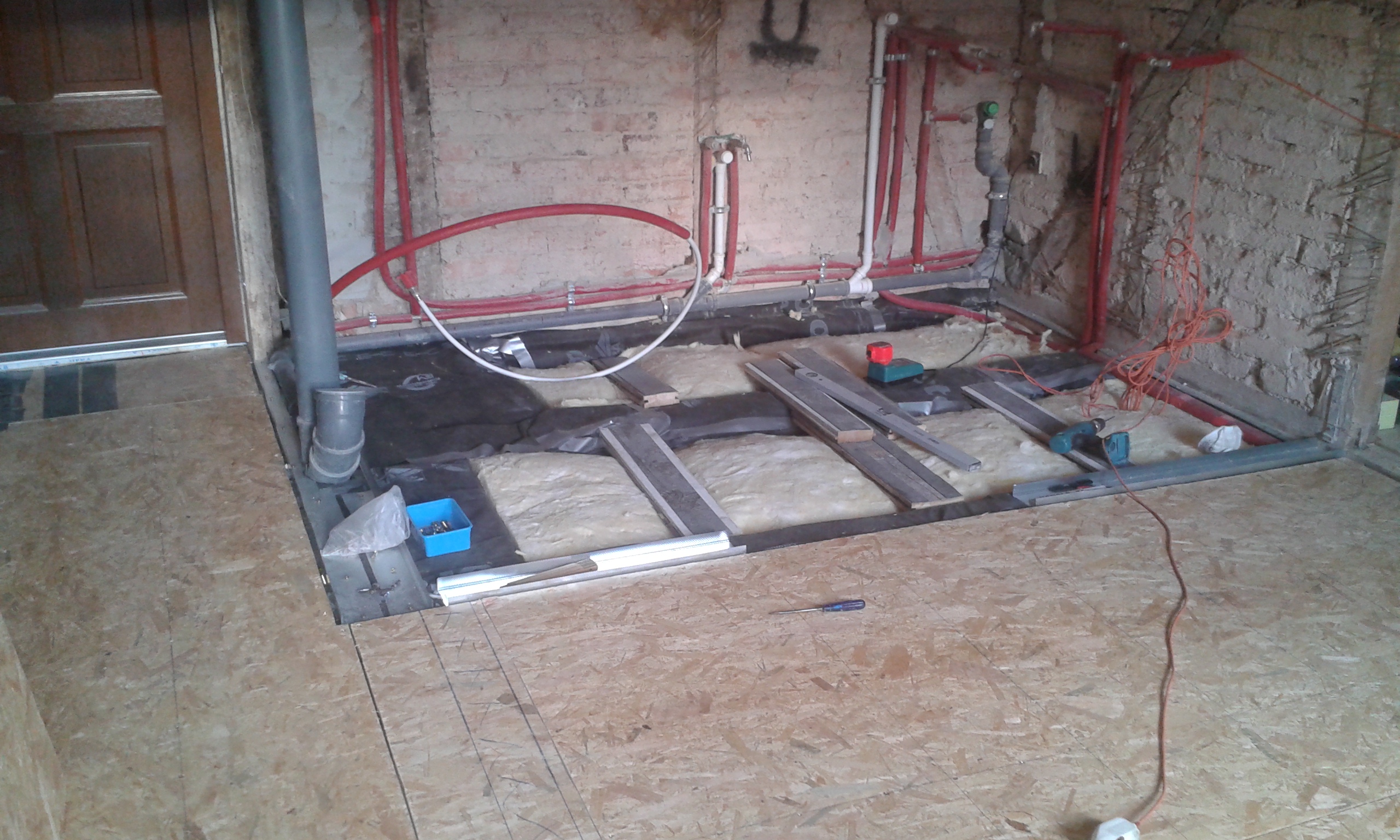 Konstrukcja podłogi w trakcie remontu – widoczne profile metalowe, izolacja termiczna oraz instalacje hydrauliczne przy ceglanej ścianie, przed zamknięciem płytami OSB.