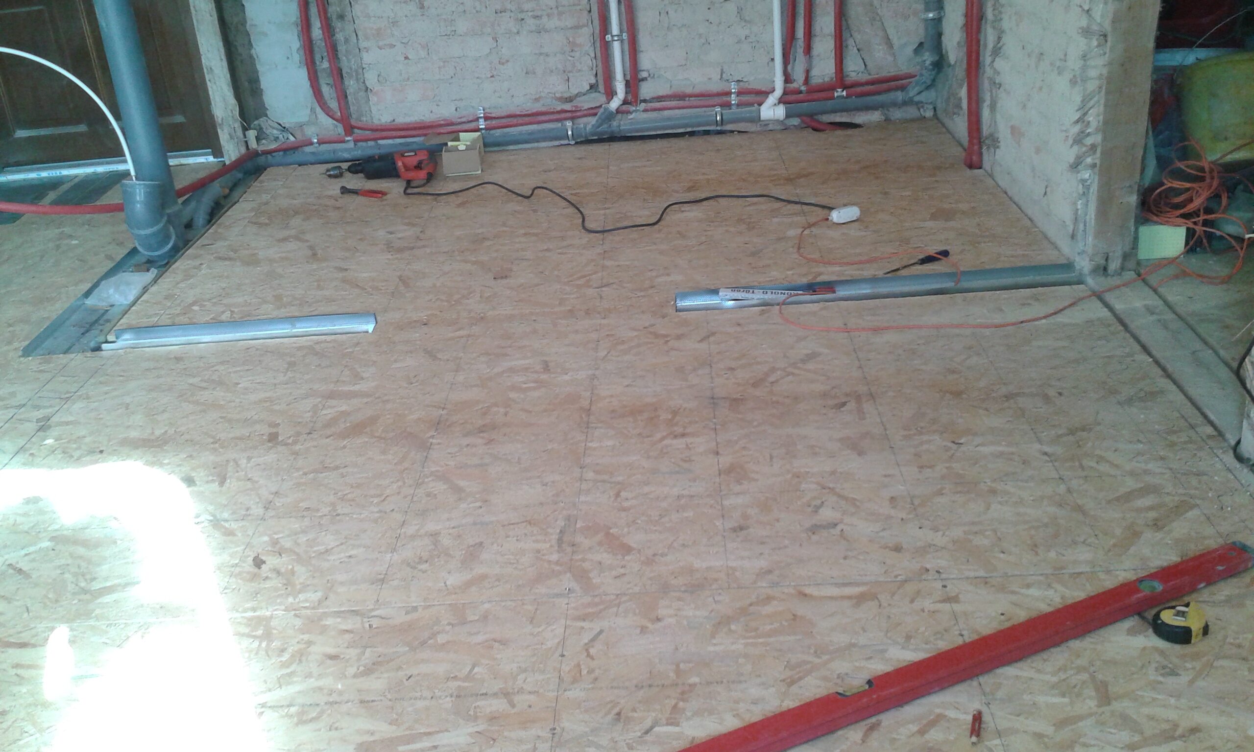 Ułożona podłoga z płyt OSB w trakcie remontu pomieszczenia, z widocznymi instalacjami hydraulicznymi przy ceglanej ścianie oraz narzędziami na podłodze.