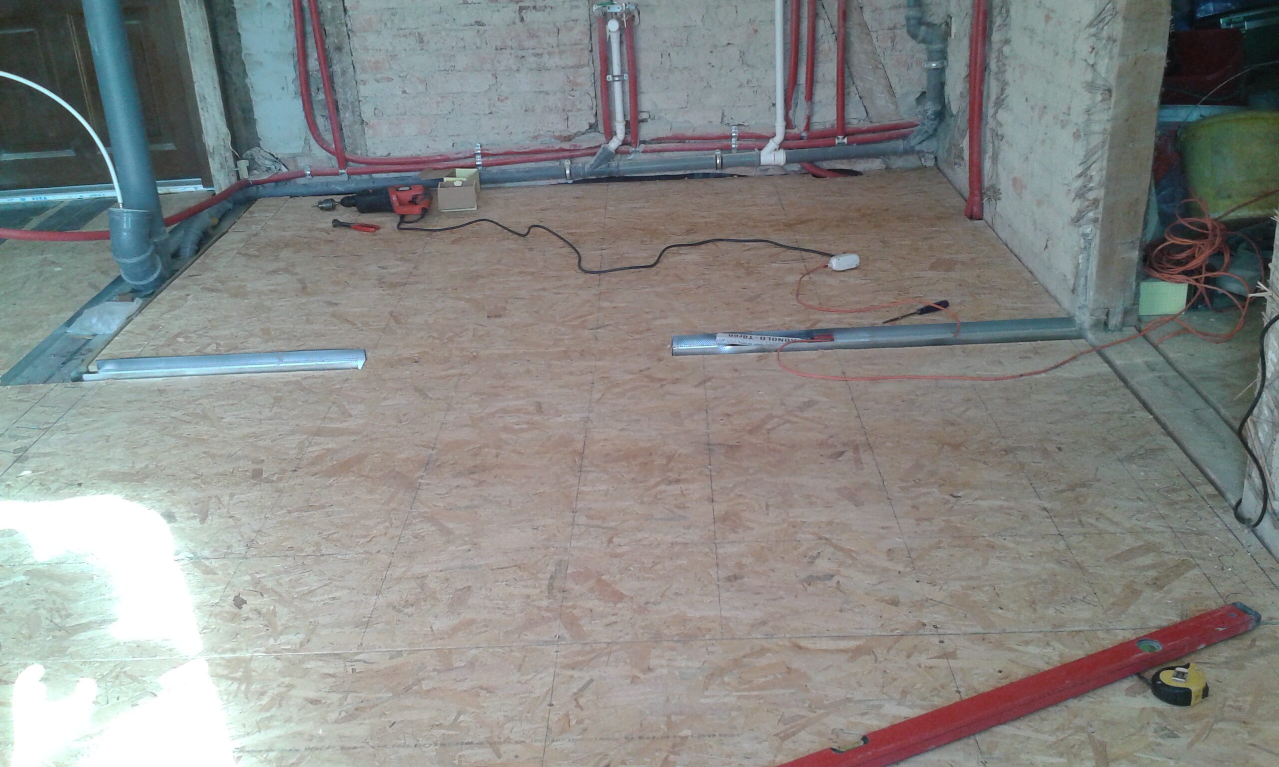 Podłoga z płyt OSB położona w pomieszczeniu podczas remontu, w tle ceglana ściana z widocznymi instalacjami hydraulicznymi i elektrycznymi.