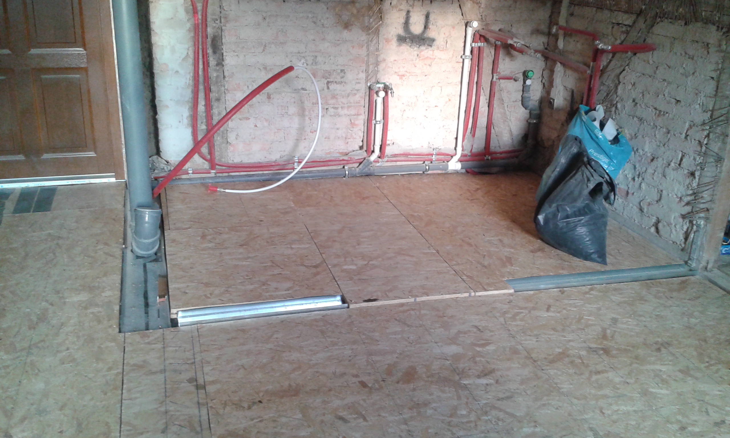 Podłoga z płyt OSB ułożonych na konstrukcji, widoczne instalacje hydrauliczne i pion kanalizacyjny przy ceglanej ścianie, wnętrze w trakcie remontu.