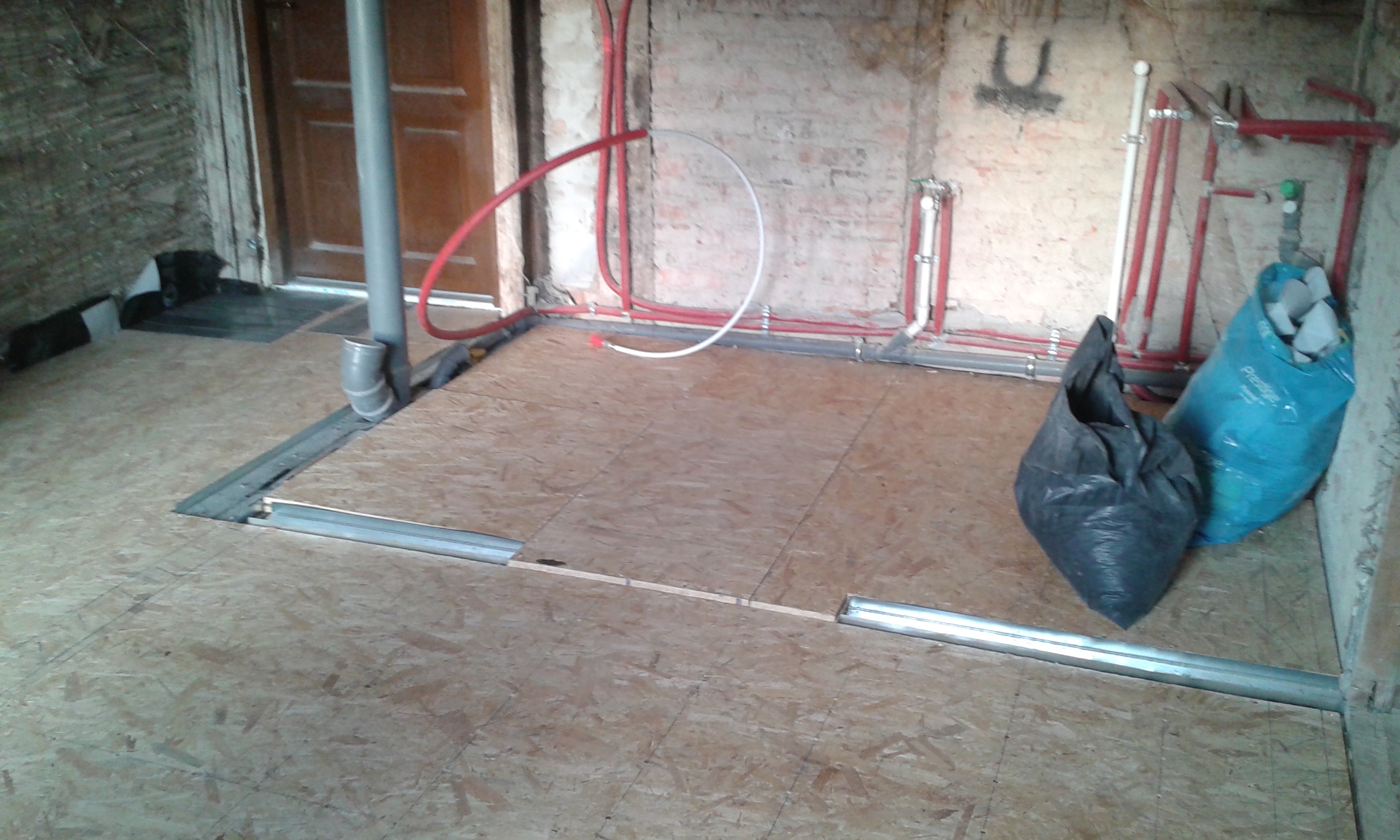 Podłoga z płyt OSB ułożonych na konstrukcji, widoczne rury instalacyjne i pion kanalizacyjny przy ceglanej ścianie, wnętrze w trakcie remontu.