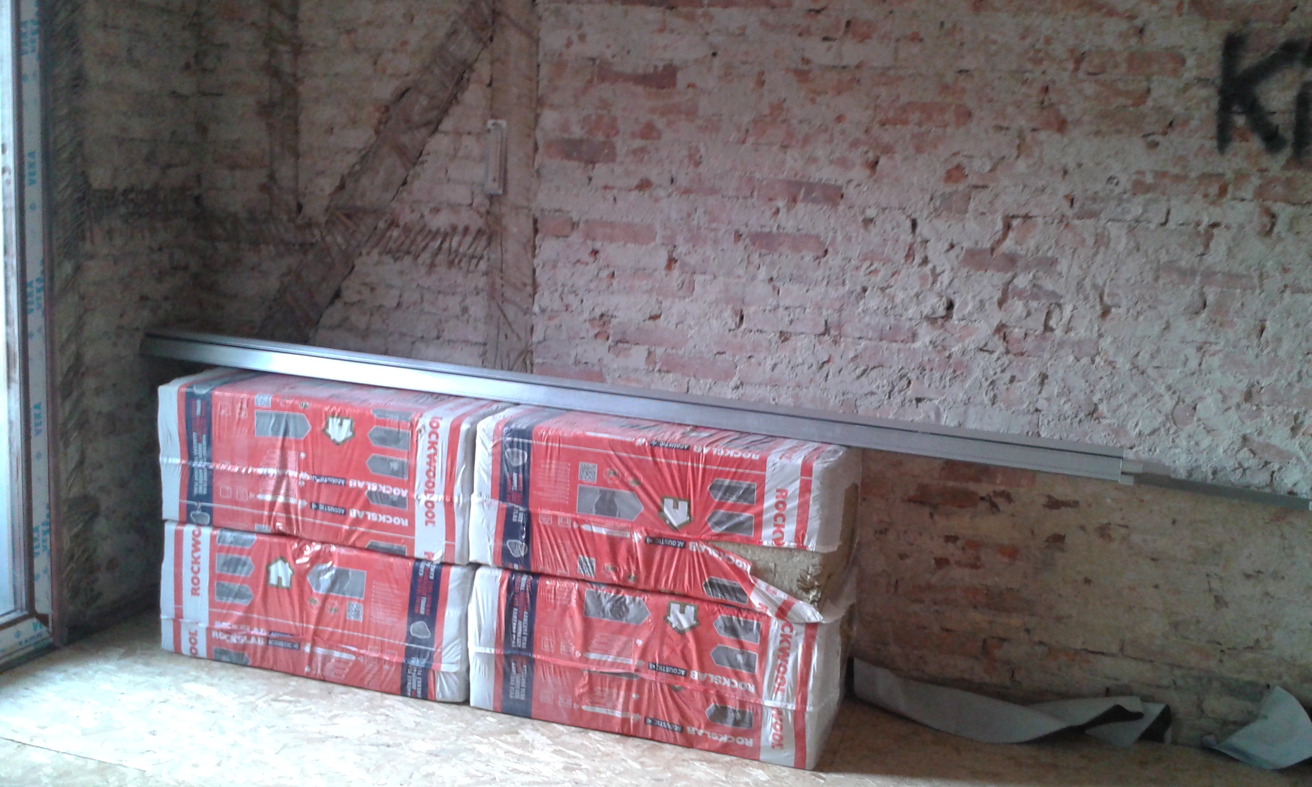 Zapakowane w folię czerwone płyty izolacyjne ułożone przy ceglanej ścianie, na nich długi metalowy profil, wnętrze w trakcie remontu i prac wykończeniowych.