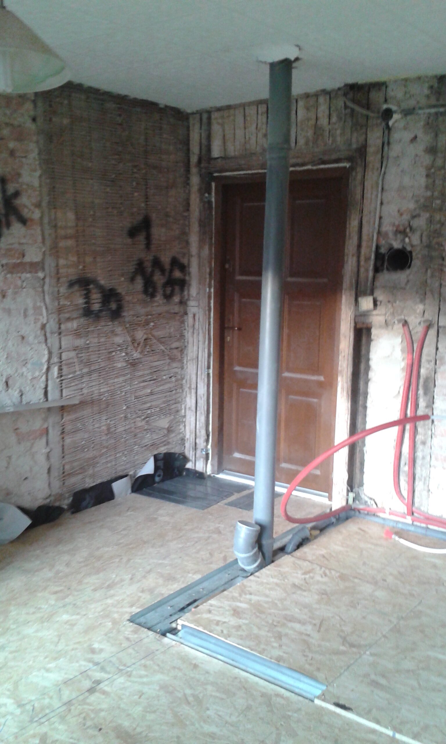 Pionowa szara rura kanalizacyjna przechodząca przez strop i podłogę z płyt OSB, prowadzone metalowe profile w podłodze, widoczne czerwone rury instalacyjne przy ścianie oraz drzwi wejściowe w stanie surowym podczas remontu.