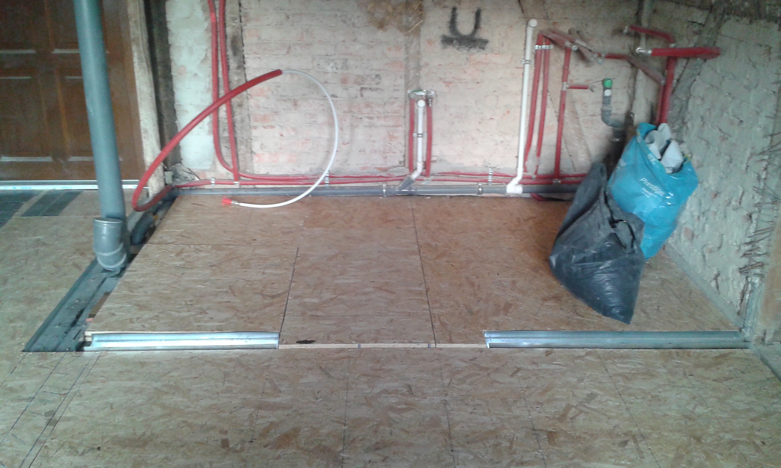 Podłoga z płyt OSB ułożonych na stelażu podczas remontu, przy ścianie prowadzone czerwone rury instalacji hydraulicznej i szara rura kanalizacyjna, metalowe profile przy krawędzi przejścia, worek z odpadami budowlanymi.