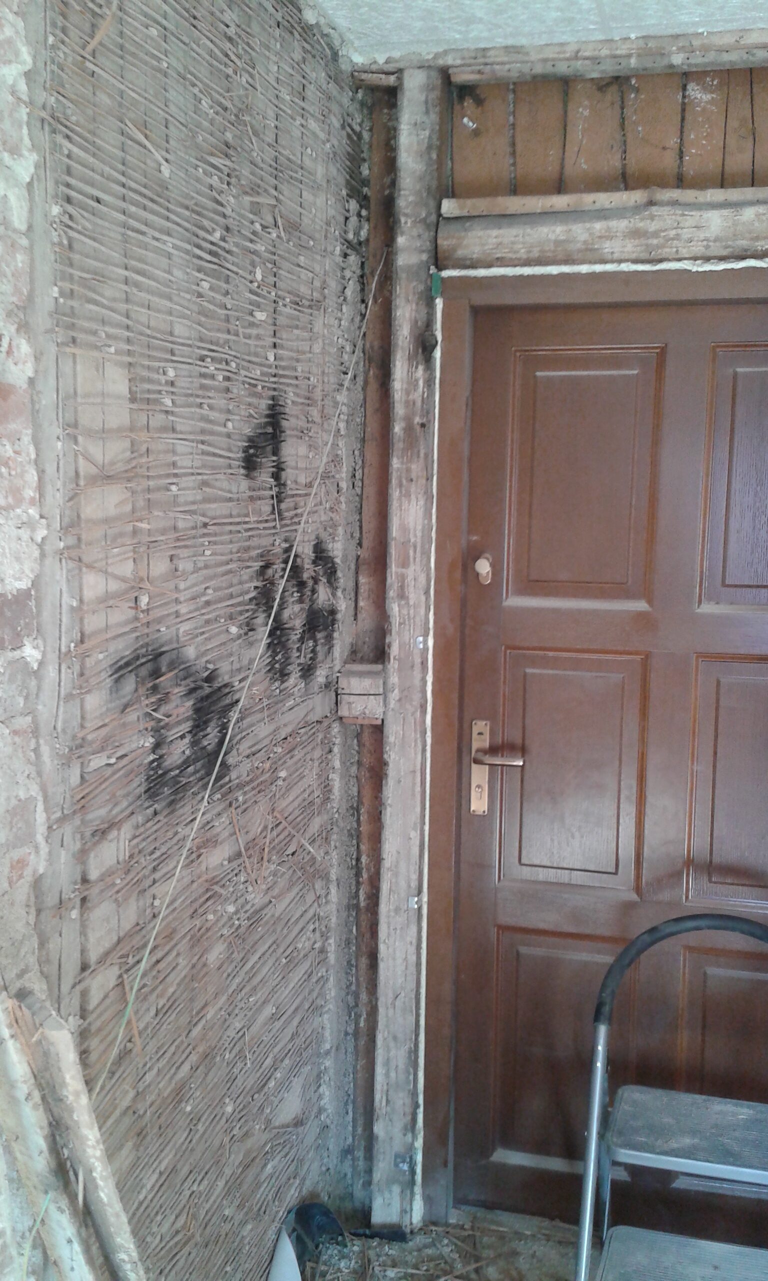 Fragment ściany przy drzwiach wejściowych z odsłoniętą konstrukcją drewnianą i matą trzcinową, widoczny podczas prac rozbiórkowych i remontowych.