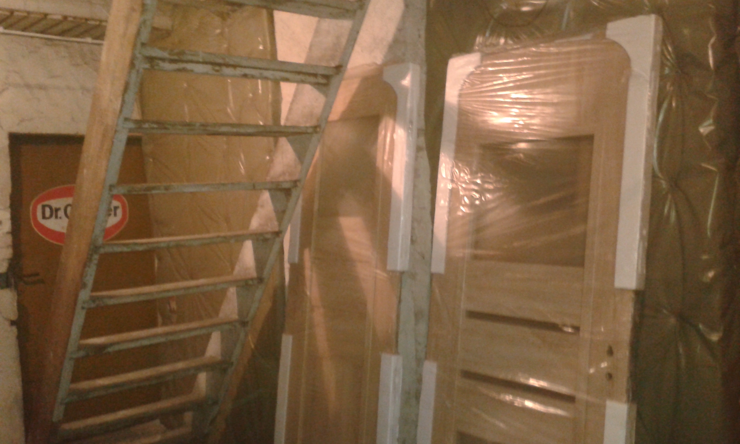 Drzwi wewnętrzne zabezpieczone folią ochronną w trakcie remontu, z widocznymi pracami budowlanymi i elementami konstrukcyjnymi klatki schodowej.