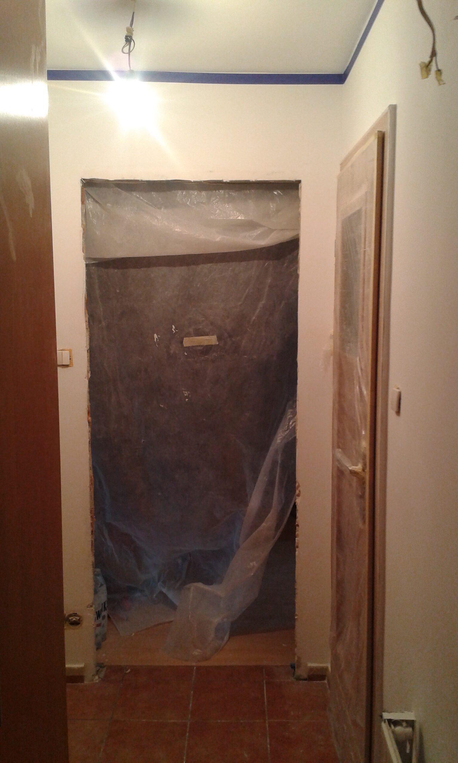 Otwór drzwiowy zabezpieczony folią ochronną podczas remontu, z widocznymi pracami wykończeniowymi i przygotowaniem ścian w korytarzu.