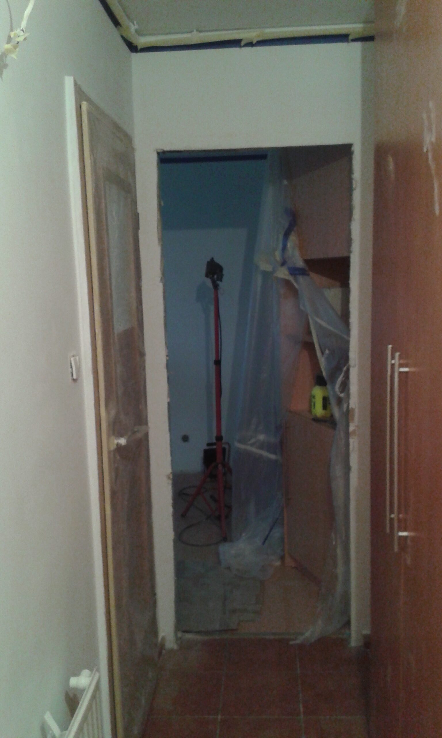 Widok przejścia między pomieszczeniami w trakcie prac remontowych, z zabezpieczoną zabudową, odsłoniętym otworem drzwiowym i sprzętem budowlanym.