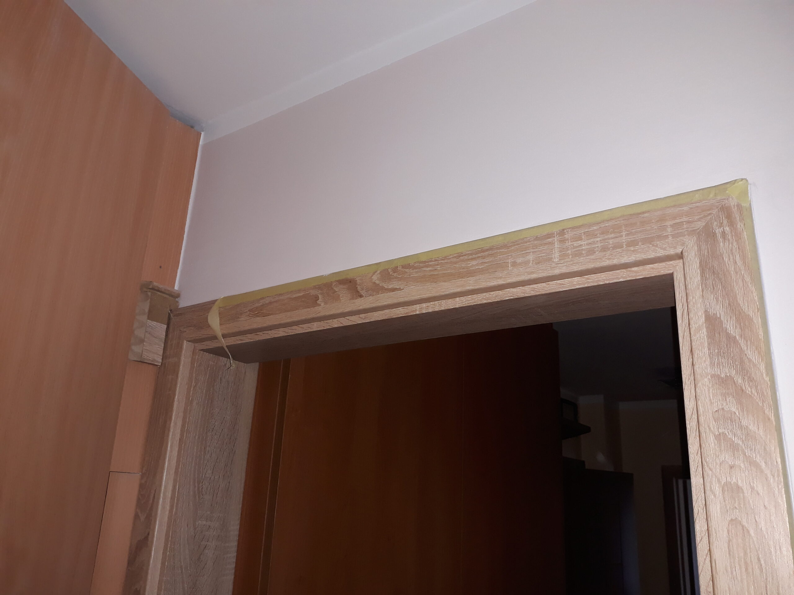 Zbliżenie na górną część ościeżnicy drzwiowej w trakcie prac wykończeniowych, z widocznym zabezpieczeniem krawędzi przed dalszym montażem.