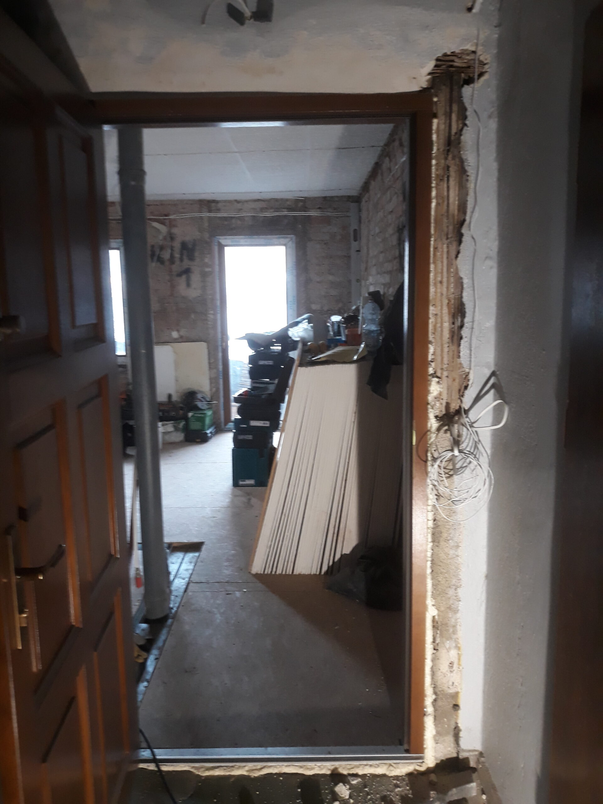 Widok przez otwór drzwiowy na wnętrze w trakcie remontu, z odsłoniętymi ścianami, materiałami budowlanymi i przygotowaną ościeżnicą.