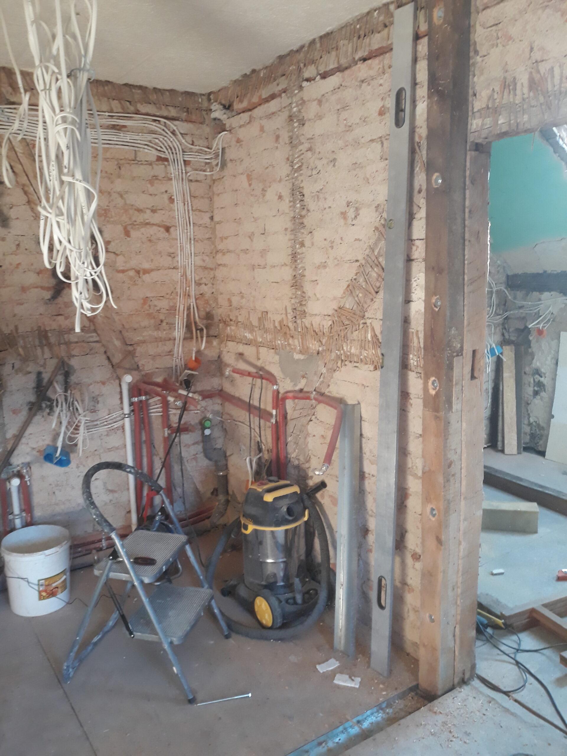 Odsłonięta ceglana ściana podczas remontu, widoczne prowadzone przewody elektryczne i rury hydrauliczne, pionowy metalowy profil przy krawędzi ściany, odkurzacz budowlany, drabinka i narzędzia ustawione na podłodze.