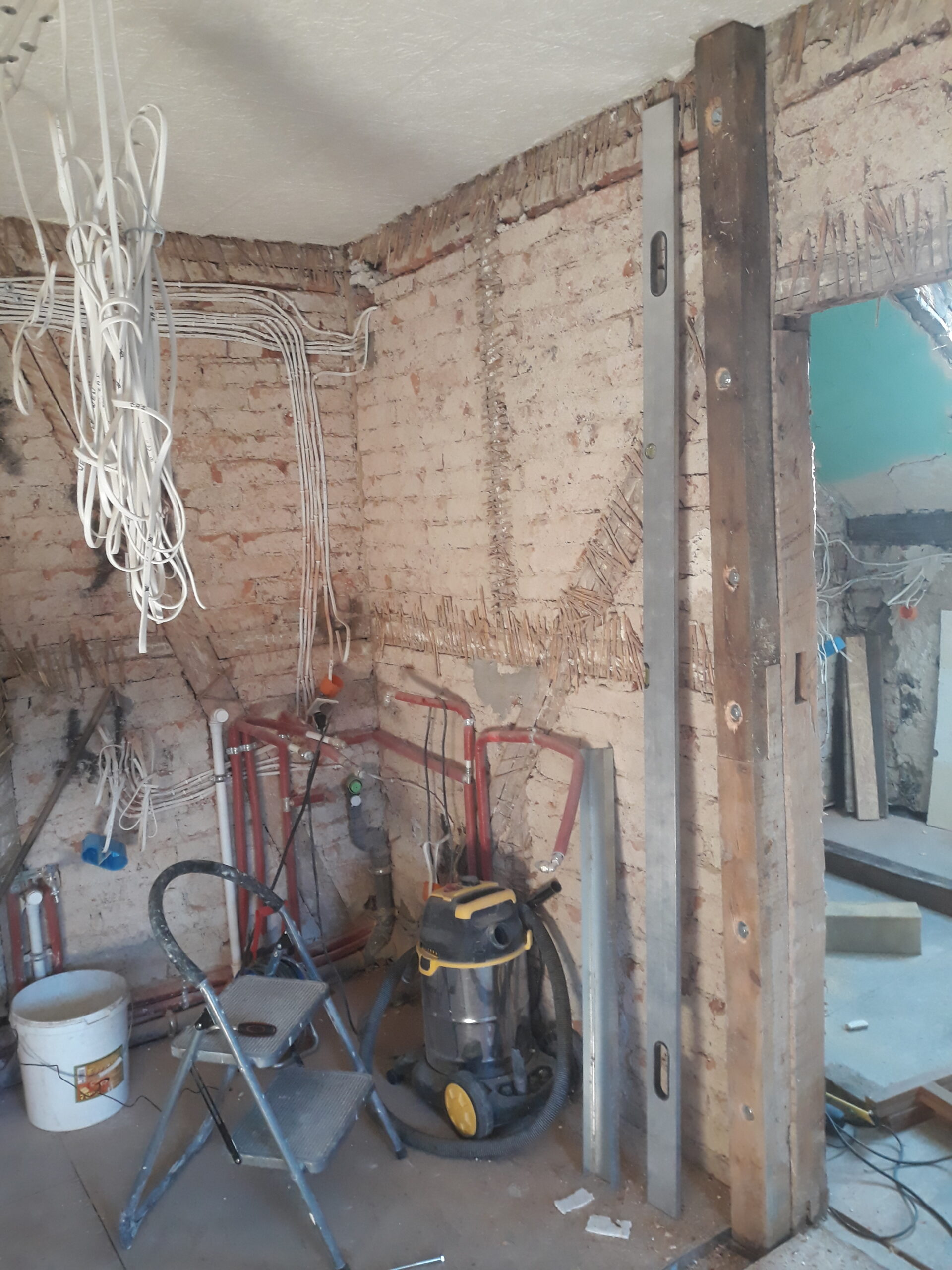 Odsłonięta ceglana ściana podczas remontu – prowadzone przewody elektryczne i rury hydrauliczne, drewniana konstrukcja ściany działowej z metalowym profilem, poziomica oparta o słup, odkurzacz budowlany i narzędzia na podłodze.