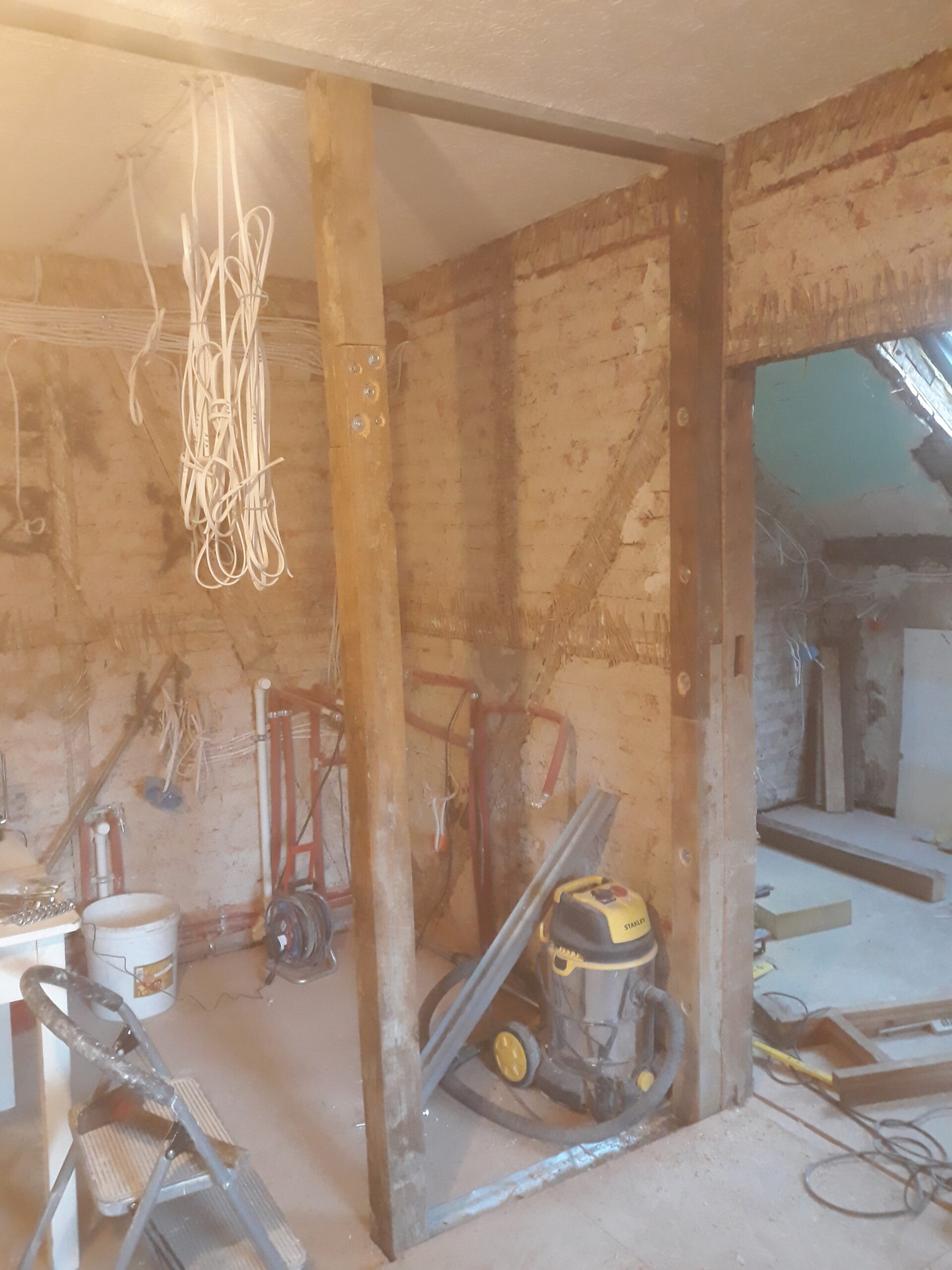 Drewniana konstrukcja ściany działowej podczas remontu – pionowe belki nośne, odsłonięta ceglana ściana z instalacją hydrauliczną, zwisające przewody elektryczne oraz odkurzacz budowlany i narzędzia na podłodze.