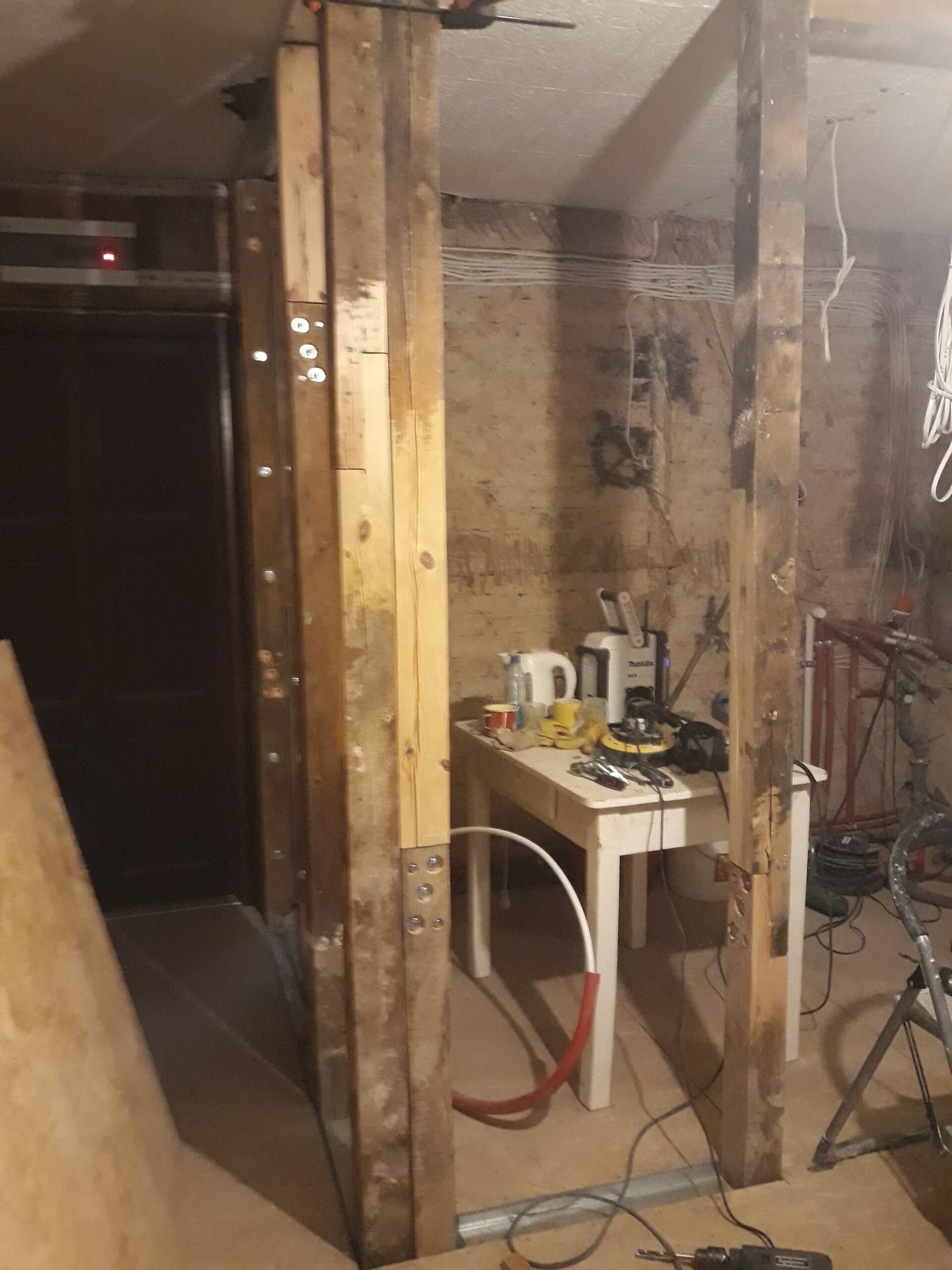 Drewniana konstrukcja ściany działowej w trakcie remontu – pionowe belki konstrukcyjne, metalowe profile przy podłodze, stół roboczy z narzędziami oraz widoczne przewody i instalacje przy ceglanej ścianie.