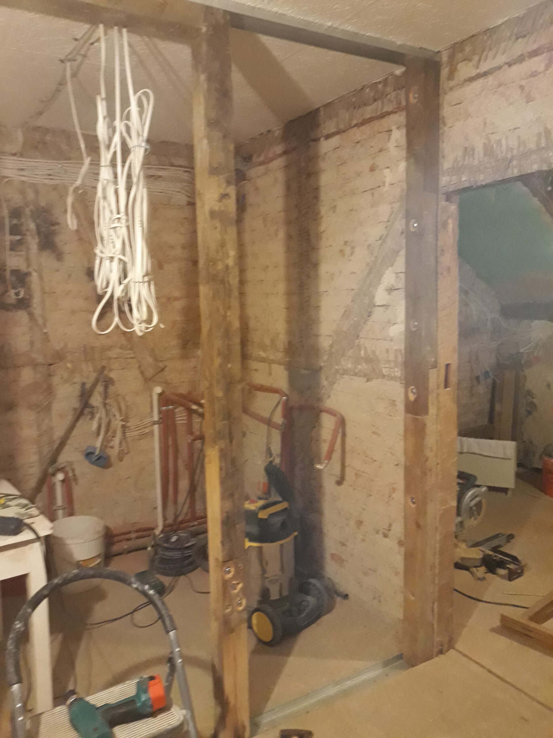 Drewniana konstrukcja ściany działowej podczas remontu – widoczne pionowe belki, metalowe profile przy podłodze i suficie, wiązka przewodów elektrycznych zwisająca z sufitu oraz elementy instalacji hydraulicznej przy ceglanej ścianie.
