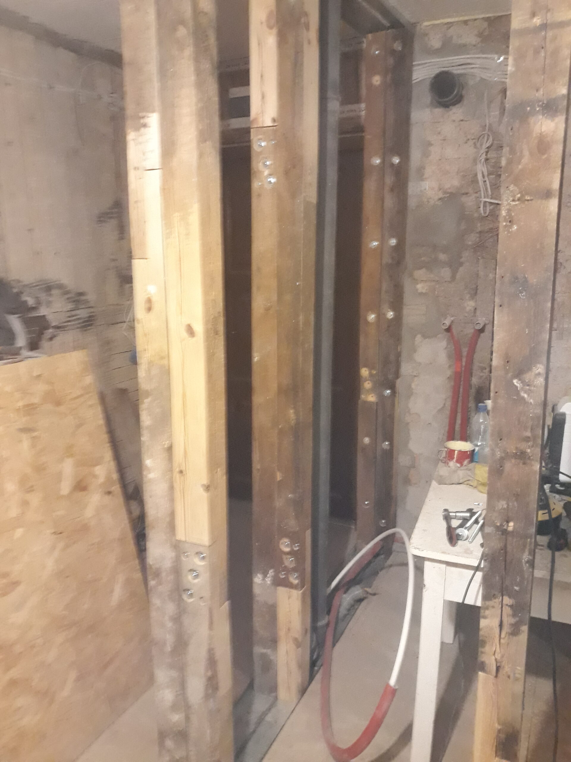 Drewniana konstrukcja ściany działowej w trakcie remontu – widoczne pionowe słupki wzmacniane metalowymi łącznikami, fragmenty instalacji wodnej i elektrycznej oraz prowizoryczny stół roboczy w tle.