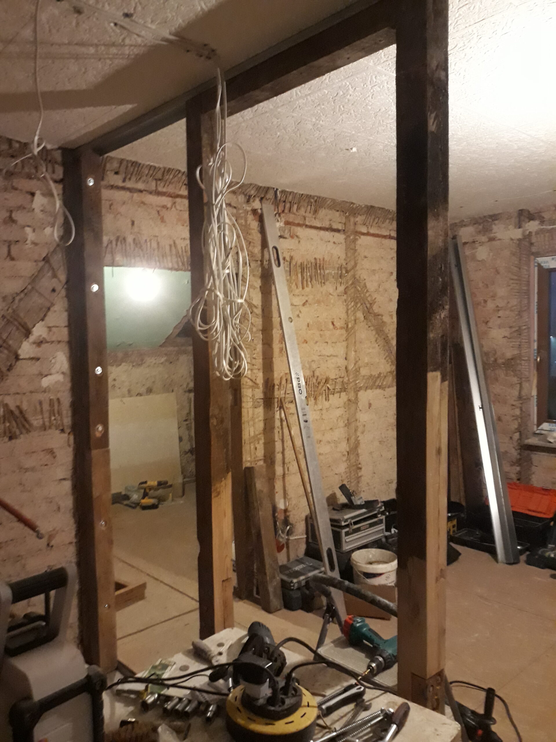 Widok wnętrza w trakcie remontu – drewniana konstrukcja ściany działowej z pionowymi słupkami, zwisającymi przewodami instalacji elektrycznej oraz odsłoniętą ceglaną ścianą w tle, na podłodze widoczne narzędzia budowlane.