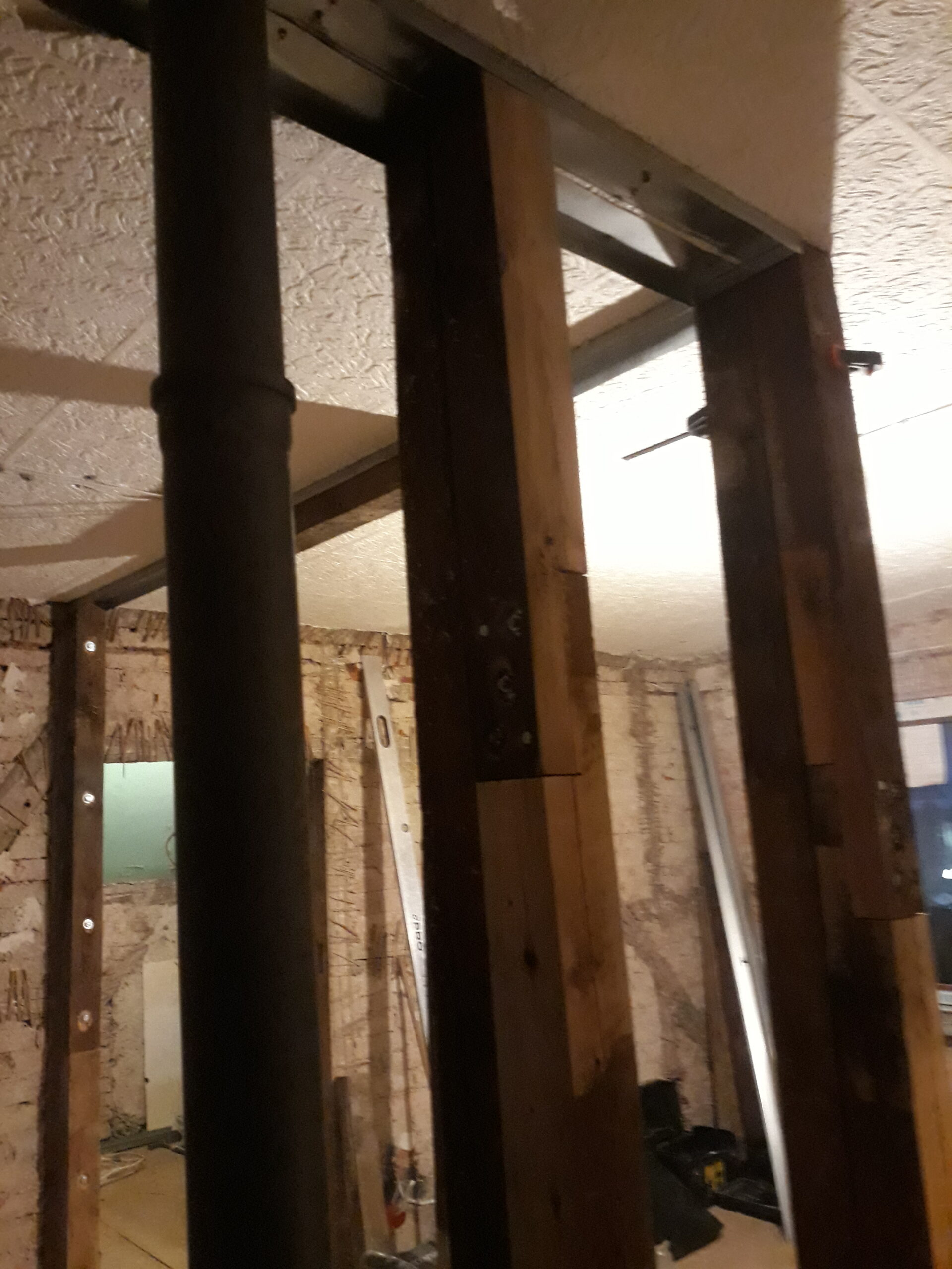 Widok górnej części konstrukcji ściany w trakcie remontu – drewniane słupki nośne połączone z metalowym profilem przy suficie, widoczna pionowa rura instalacyjna oraz surowe ściany ceglane w tle.