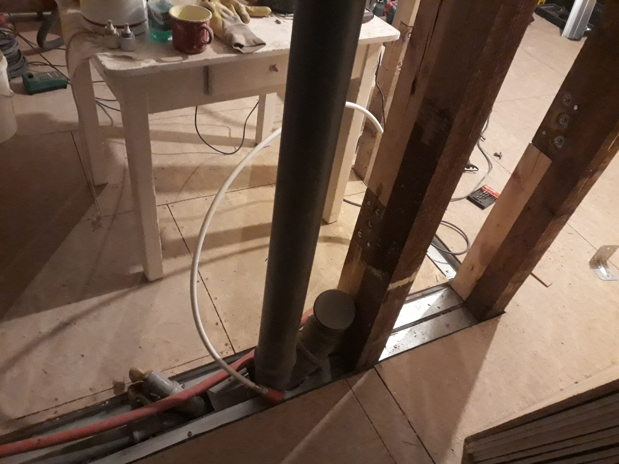 Zbliżenie na podłogę w trakcie remontu – pionowa rura kanalizacyjna, przewody instalacyjne oraz drewniane słupki konstrukcyjne ściany, widoczne elementy montażowe i instalacje prowadzone w podłodze.