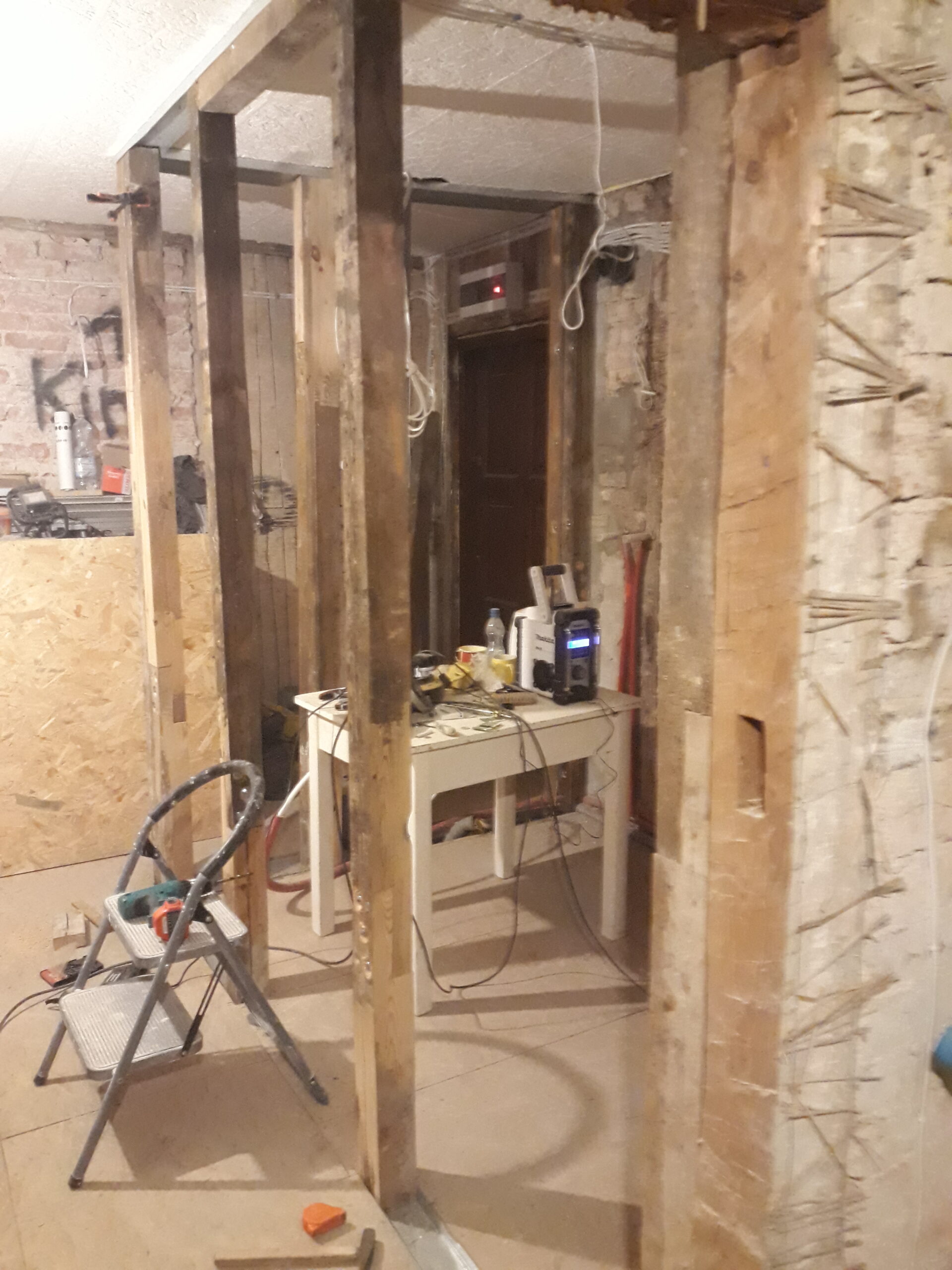 Remont poddasza – wnętrze w trakcie prac budowlanych z odsłoniętą drewnianą konstrukcją ścian nośnych, widoczne przewody elektryczne, prowizoryczny stół roboczy z narzędziami, składane krzesło oraz przejście do korytarza.