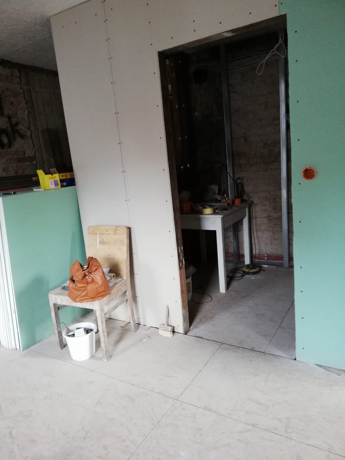 Remont poddasza – wnętrze z zabudową z płyt gipsowo-kartonowych, widoczny otwór drzwiowy do pomieszczenia technicznego, prowizoryczne krzesło z torebką, wiadro, płyty budowlane oraz fragmenty instalacji w trakcie prac wykończeniowych.