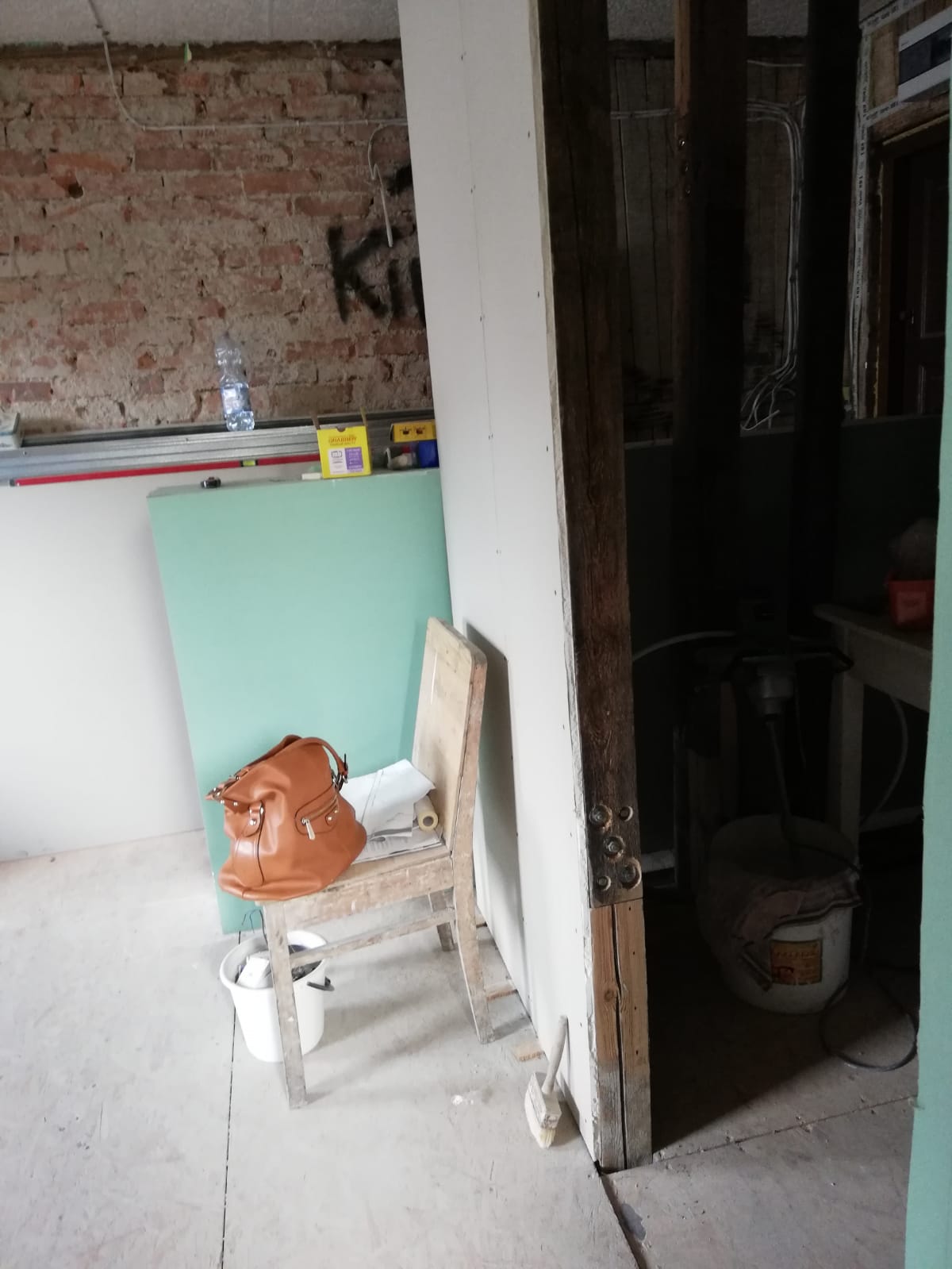 Remont poddasza – wnętrze z odsłoniętą ceglaną ścianą, fragmentem zabudowy z płyt gipsowo-kartonowych oraz wejściem do wydzielonego pomieszczenia, na pierwszym planie drewniane krzesło z torebką, płyty budowlane i narzędzia.