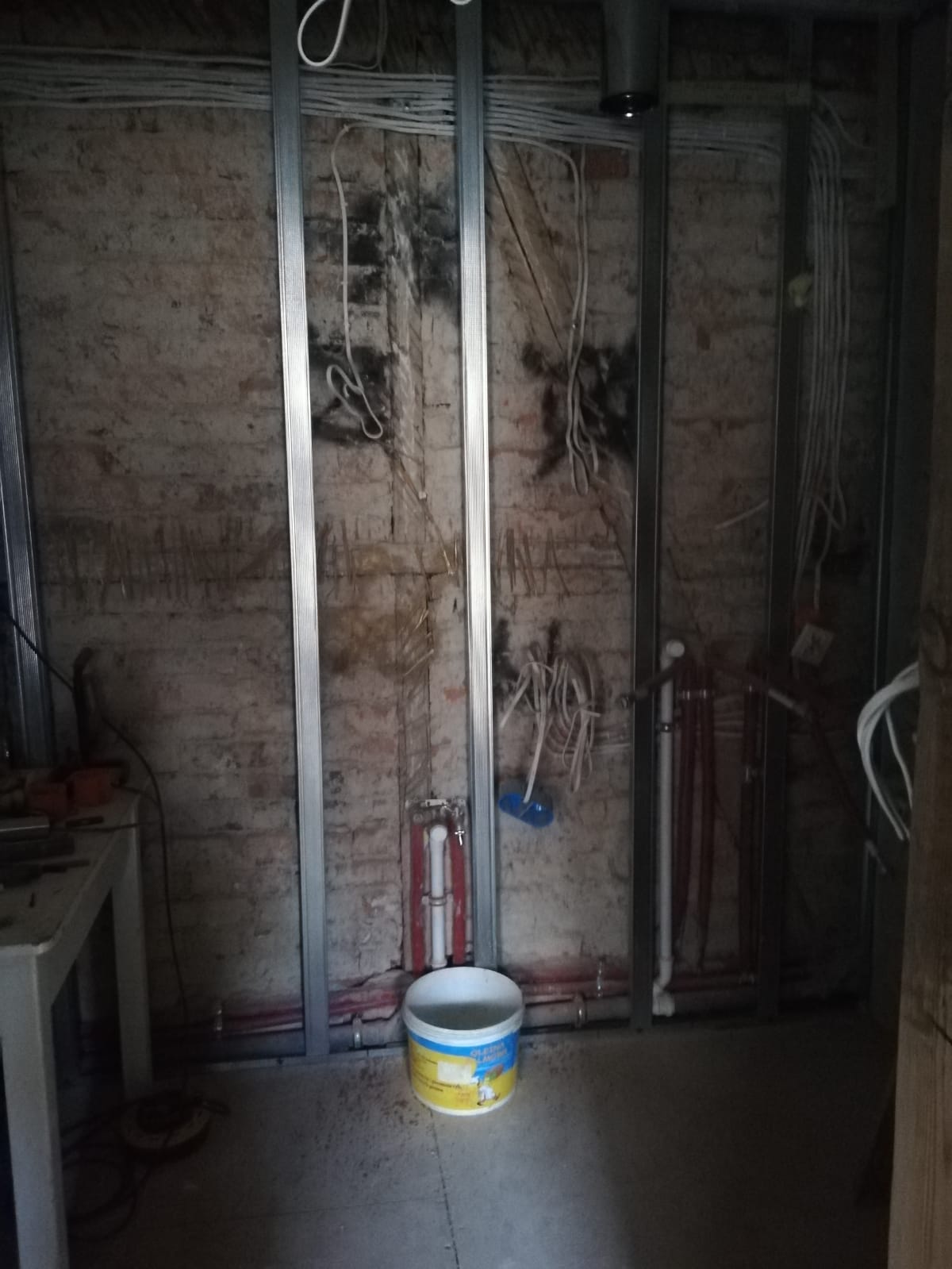 Ceglana ściana z zamontowanym metalowym stelażem, poprowadzoną instalacją elektryczną i hydrauliczną oraz przewodami przygotowanymi do dalszych prac wykończeniowych.