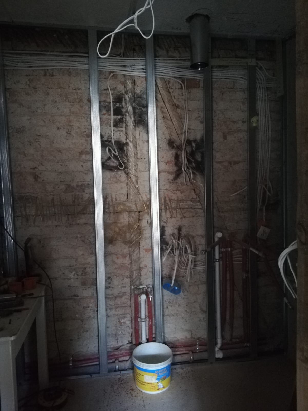Odsłonięta ceglana ściana z metalowym stelażem, poprowadzoną instalacją elektryczną i hydrauliczną oraz wiązkami przewodów w trakcie remontu wnętrza.