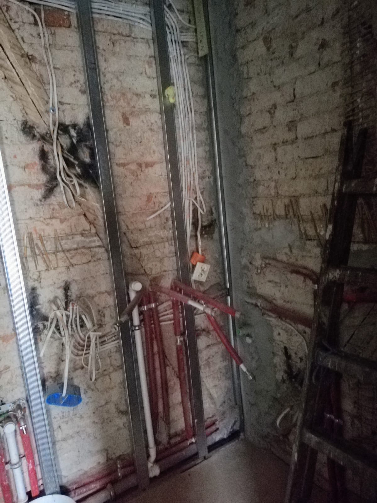 Ceglana ściana z metalowym stelażem oraz poprowadzoną instalacją elektryczną i hydrauliczną w narożniku pomieszczenia podczas remontu.