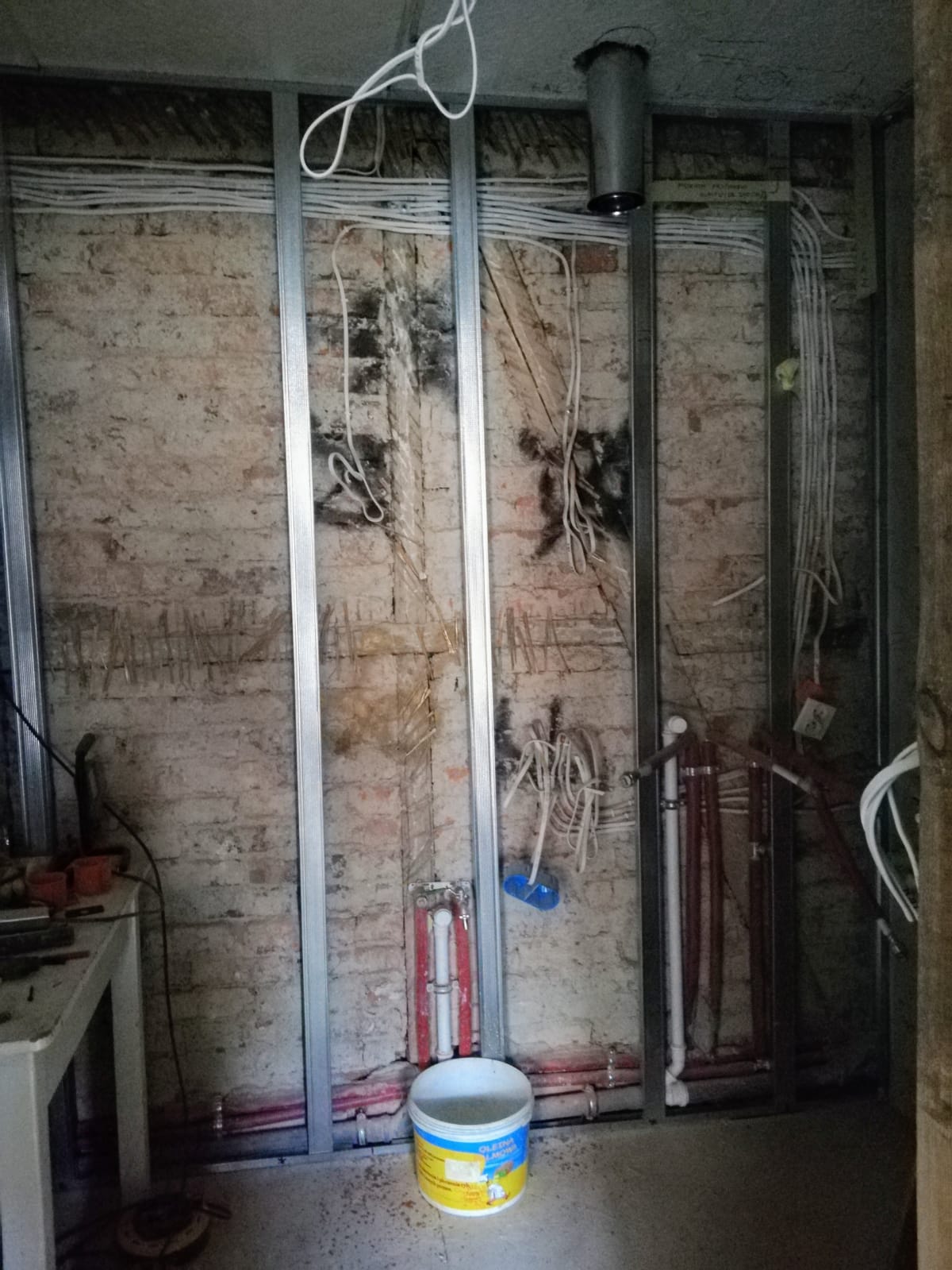 Odsłonięta ceglana ściana z metalowym stelażem oraz widoczną instalacją elektryczną i hydrauliczną w trakcie remontu.