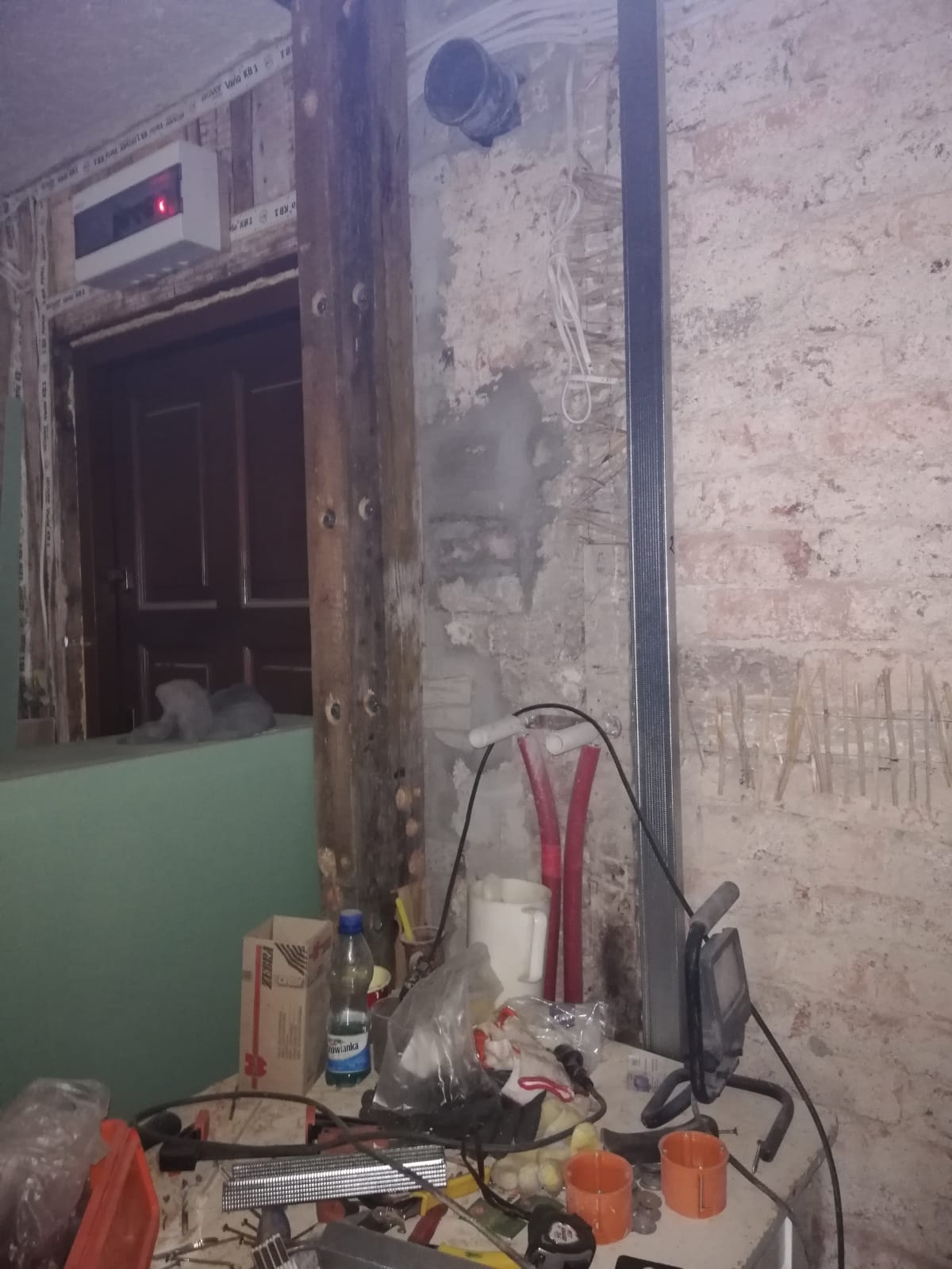 Fragment ściany w trakcie remontu z odsłoniętą cegłą, instalacją elektryczną i hydrauliczną oraz prowizorycznym stanowiskiem roboczym