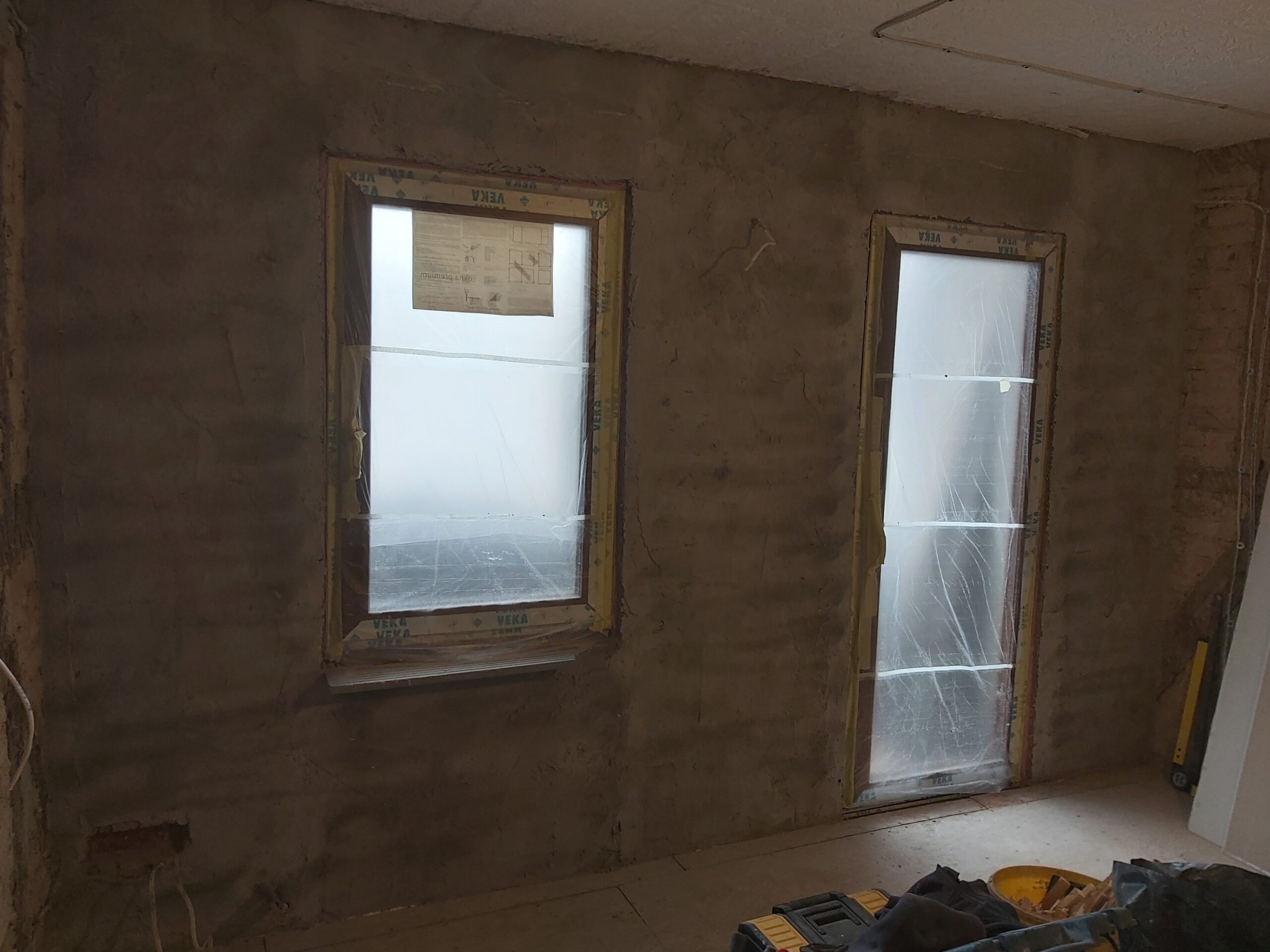 Ściana w trakcie remontu z dwoma oknami zabezpieczonymi folią ochronną, przygotowana do dalszych prac wykończeniowych.