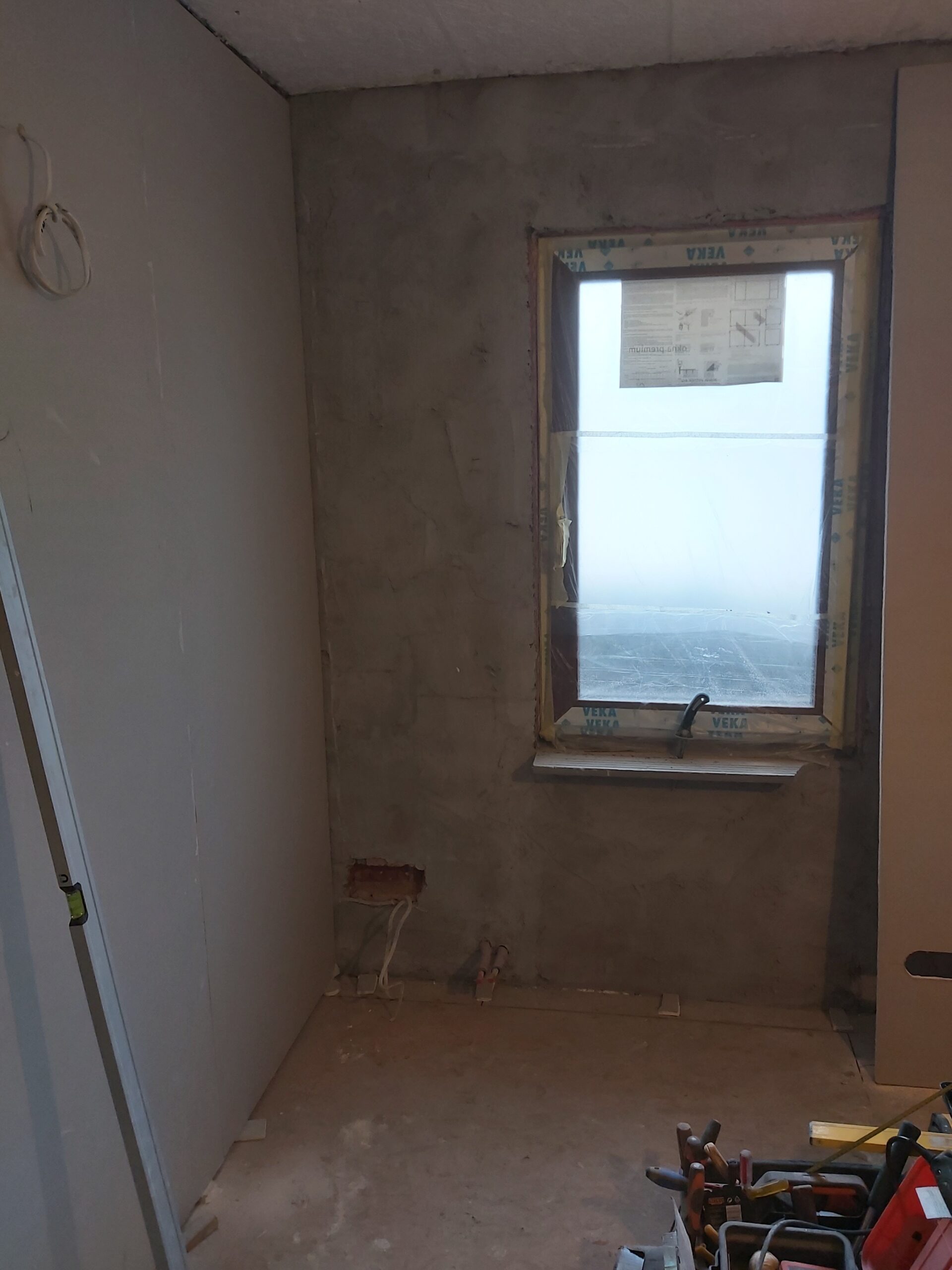 Pomieszczenie w trakcie remontu z widoczną ścianą przygotowaną pod wykończenie oraz oknem zabezpieczonym folią ochronną.