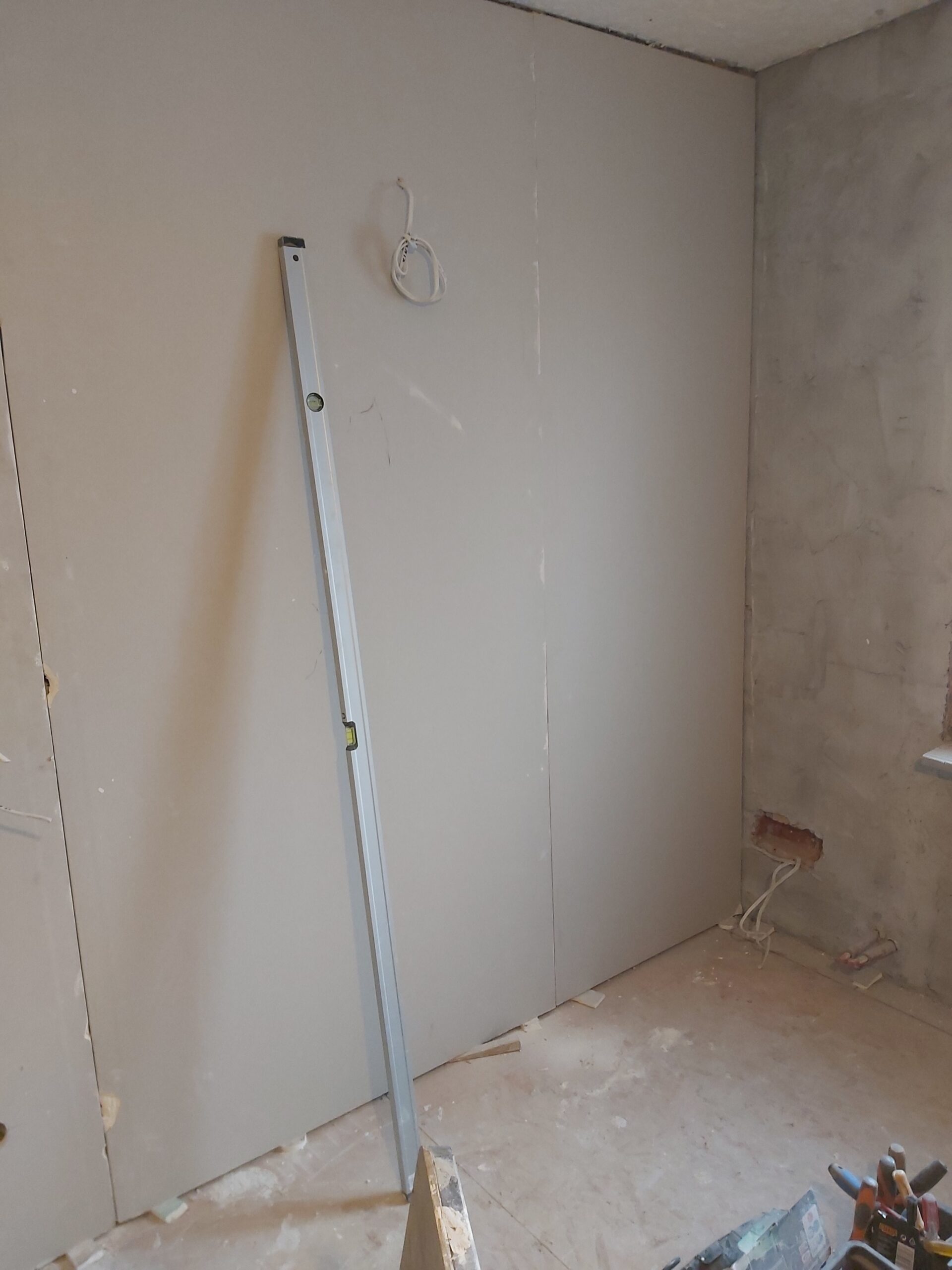 Ściana z płyt gipsowo-kartonowych w trakcie prac remontowych, widoczna niezabudowana instalacja elektryczna oraz poziomica oparta o ścianę.