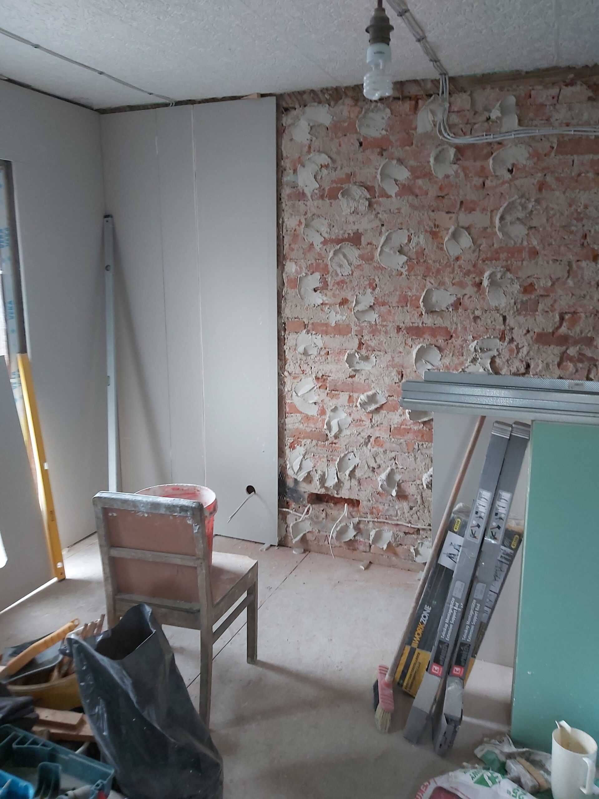 Remont poddasza – wnętrze z odsłoniętą ceglaną ścianą z widocznymi śladami po tynku, obok fragment zabudowy z płyt gipsowo-kartonowych, na podłodze krzesło, narzędzia, profile metalowe i materiały budowlane.