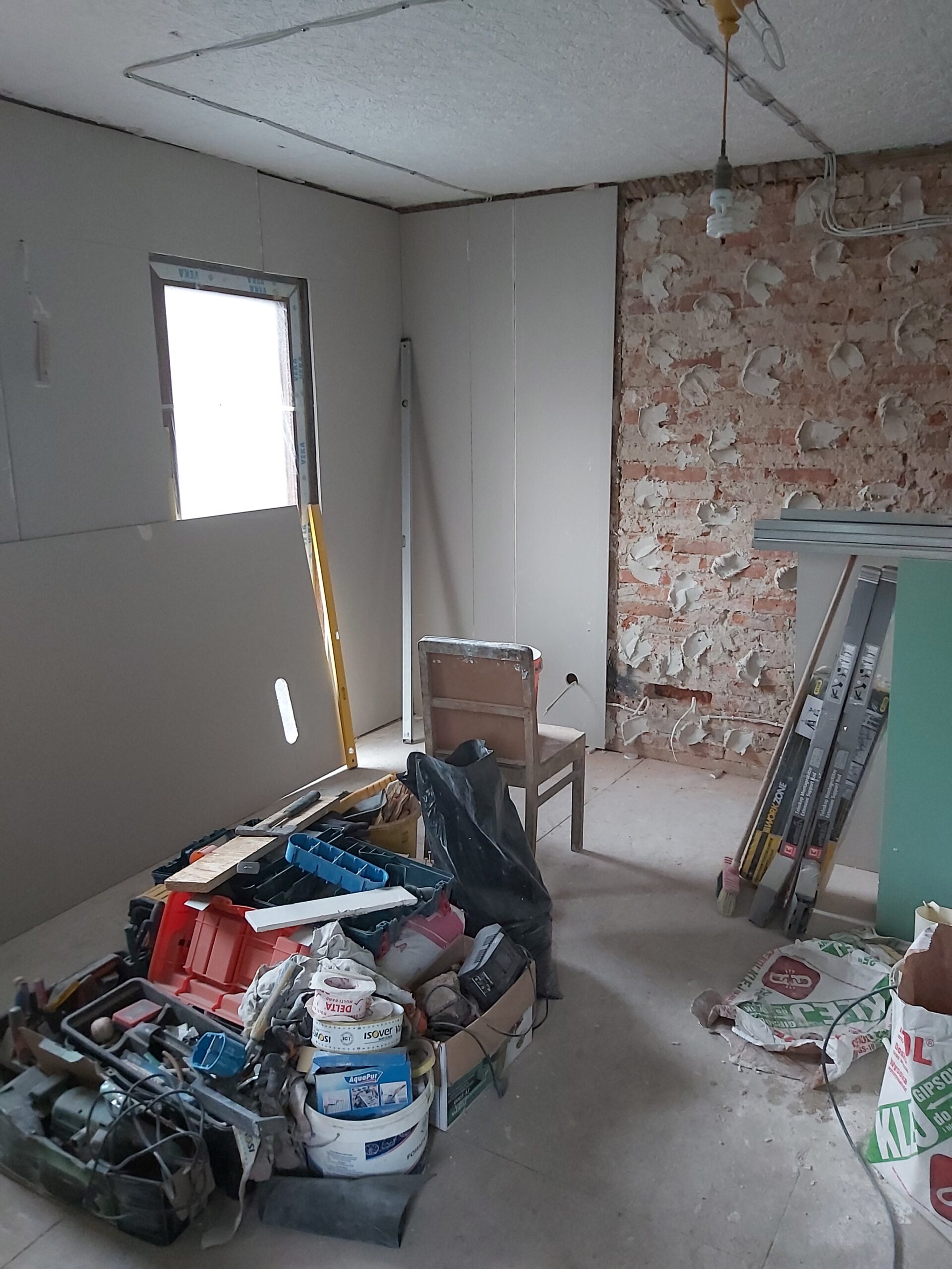 Remont poddasza – pomieszczenie z częściowo wykończonymi ścianami z płyt gipsowo-kartonowych oraz odsłoniętą ceglaną ścianą, widoczne narzędzia budowlane, skrzynki, wiadro z materiałami, krzesło i listwy montażowe.