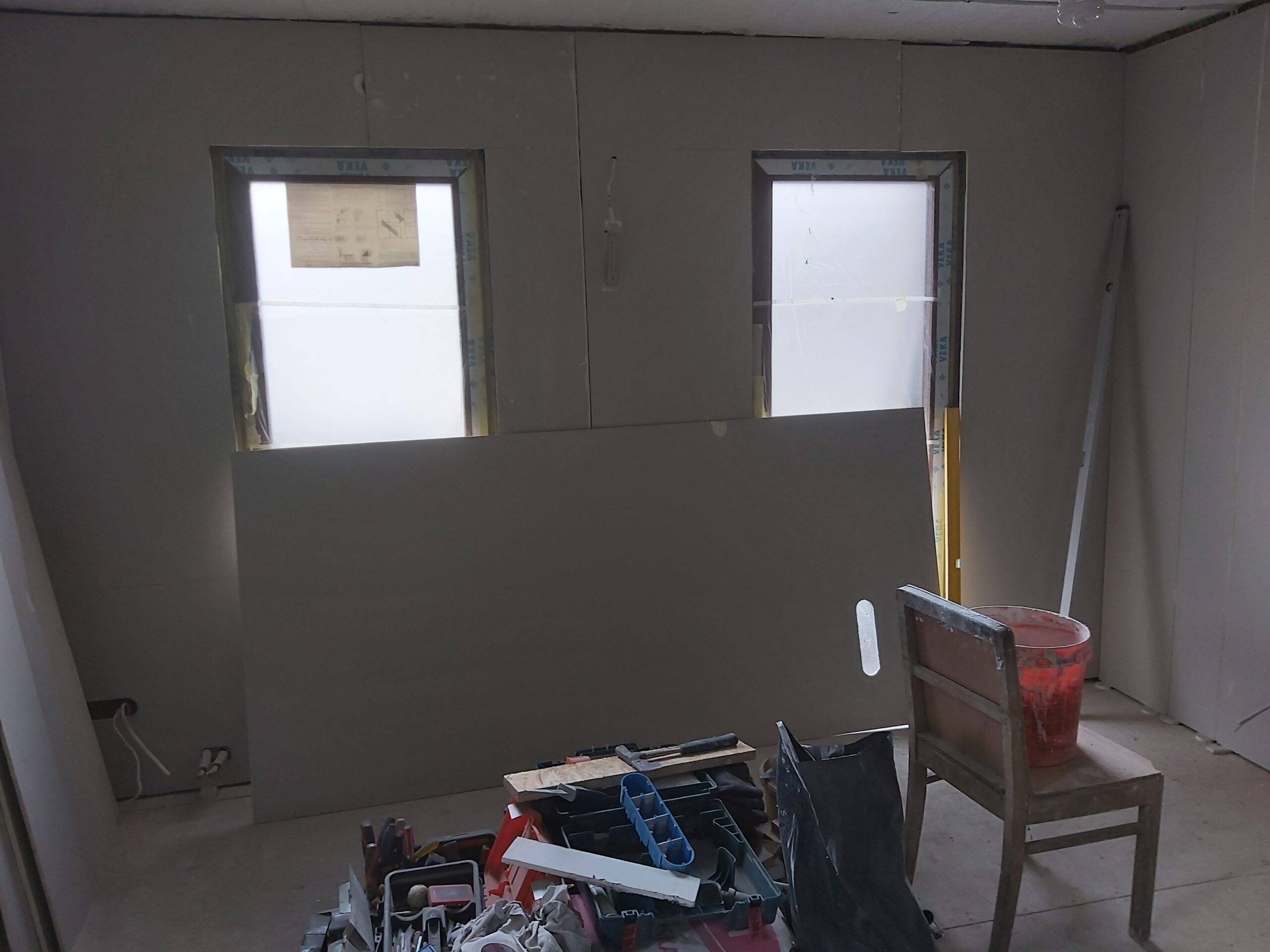 Remont poddasza – pomieszczenie z dwiema małymi oknami, ściany wykończone płytami gipsowo-kartonowymi, na pierwszym planie narzędzia, skrzynka robocza, krzesło oraz płyta GK przygotowana do montażu.