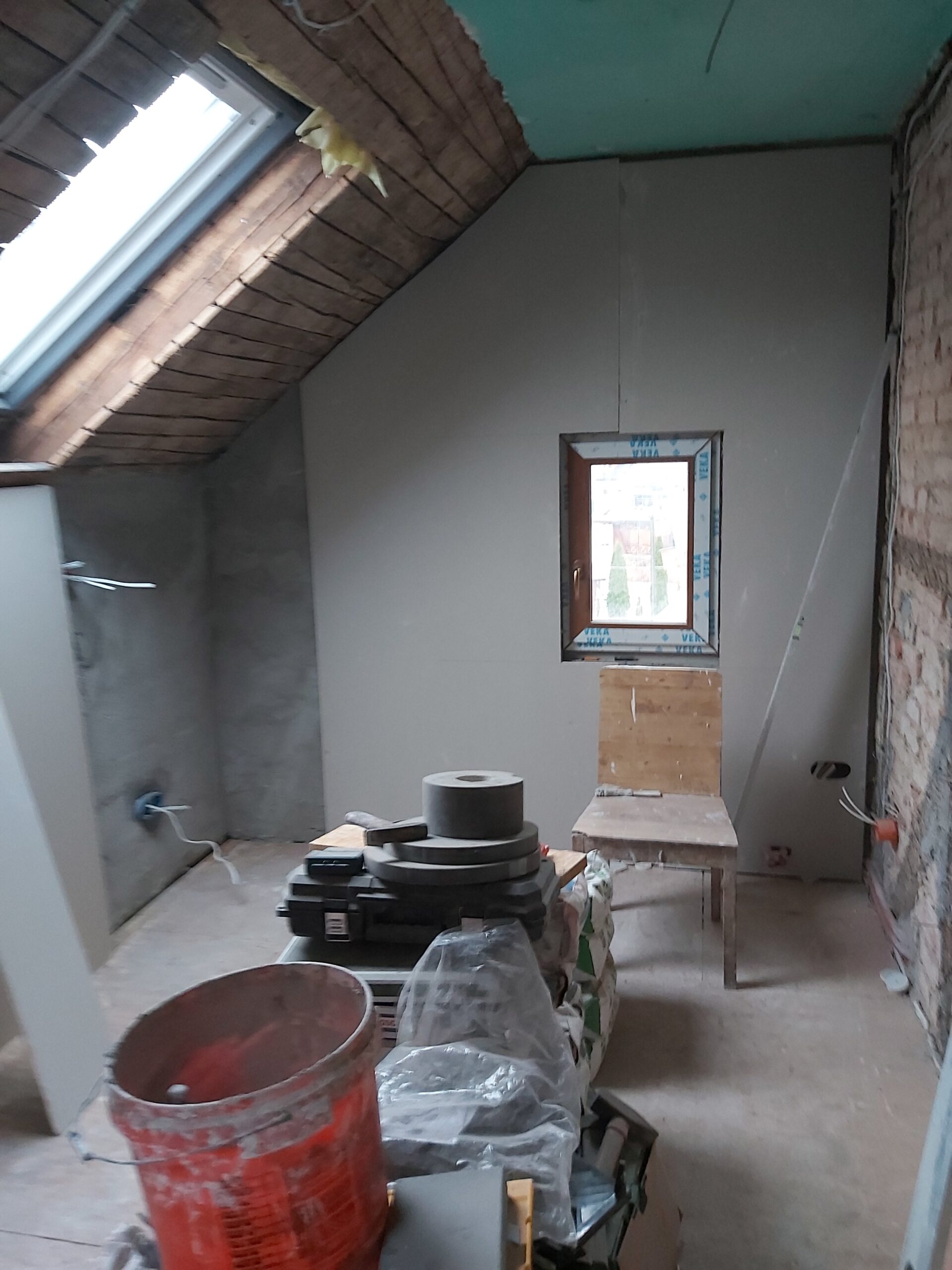 Remont poddasza – pomieszczenie z oknem dachowym i pionowym, ściany wykonane z płyt gipsowo-kartonowych, widoczne narzędzia, wiadro budowlane i materiały remontowe na środku pomieszczenia.