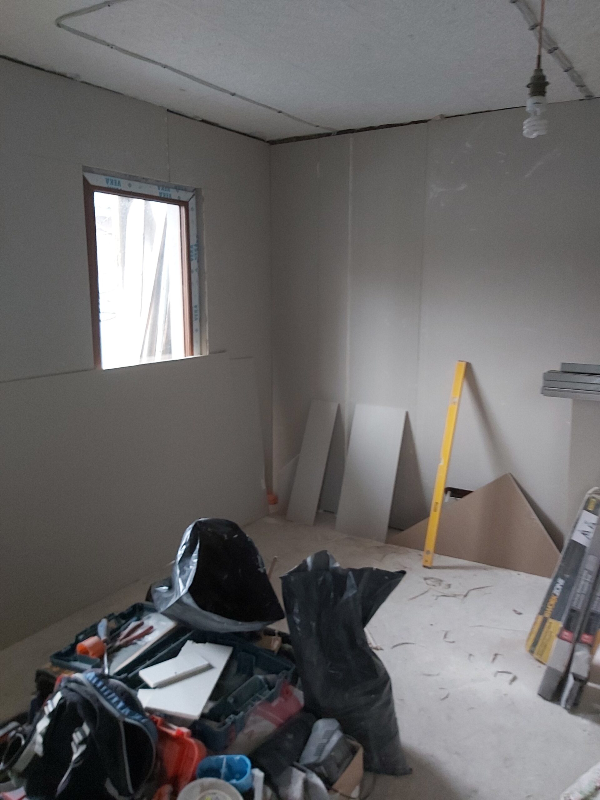 Remont poddasza – niewielki pokój z oknem, świeżo wykonane ściany z płyt gipsowo-kartonowych, na podłodze narzędzia, odpady budowlane oraz poziomnica oparta o ścianę.
