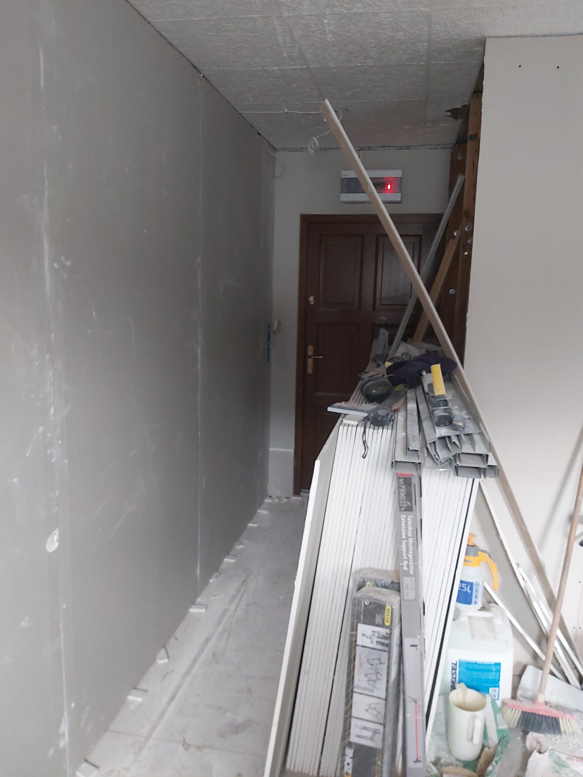 Remont poddasza – wąski korytarz z nowo wykonanymi ścianami z płyt gipsowo-kartonowych, składowane profile, płyty GK oraz narzędzia budowlane przed drzwiami wejściowymi.
