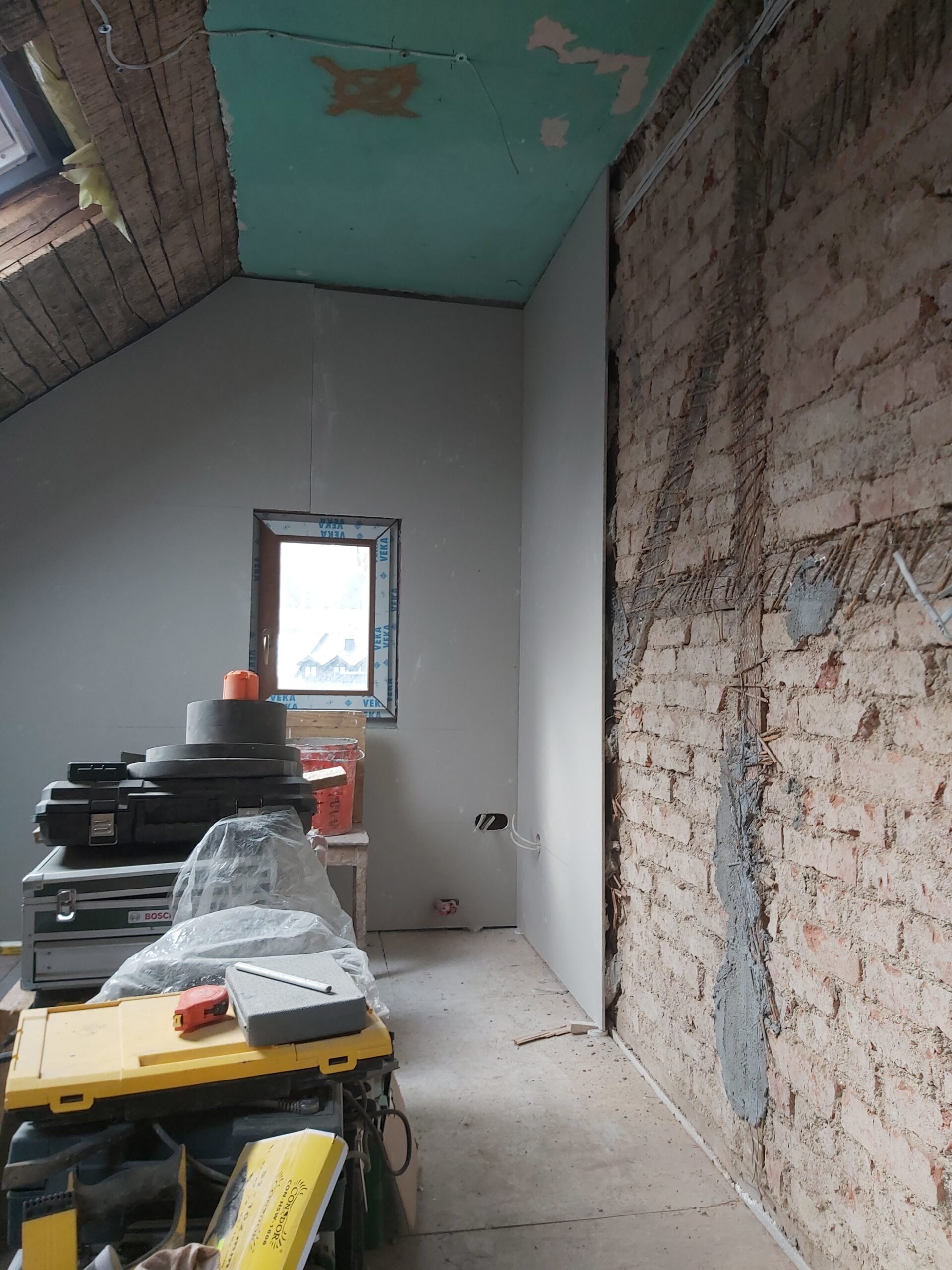 Remont poddasza – widok na ścianę z płyt gipsowo-kartonowych, odsłoniętą ceglaną ścianę nośną, okno dachowe oraz narzędzia i materiały budowlane ustawione w pomieszczeniu.