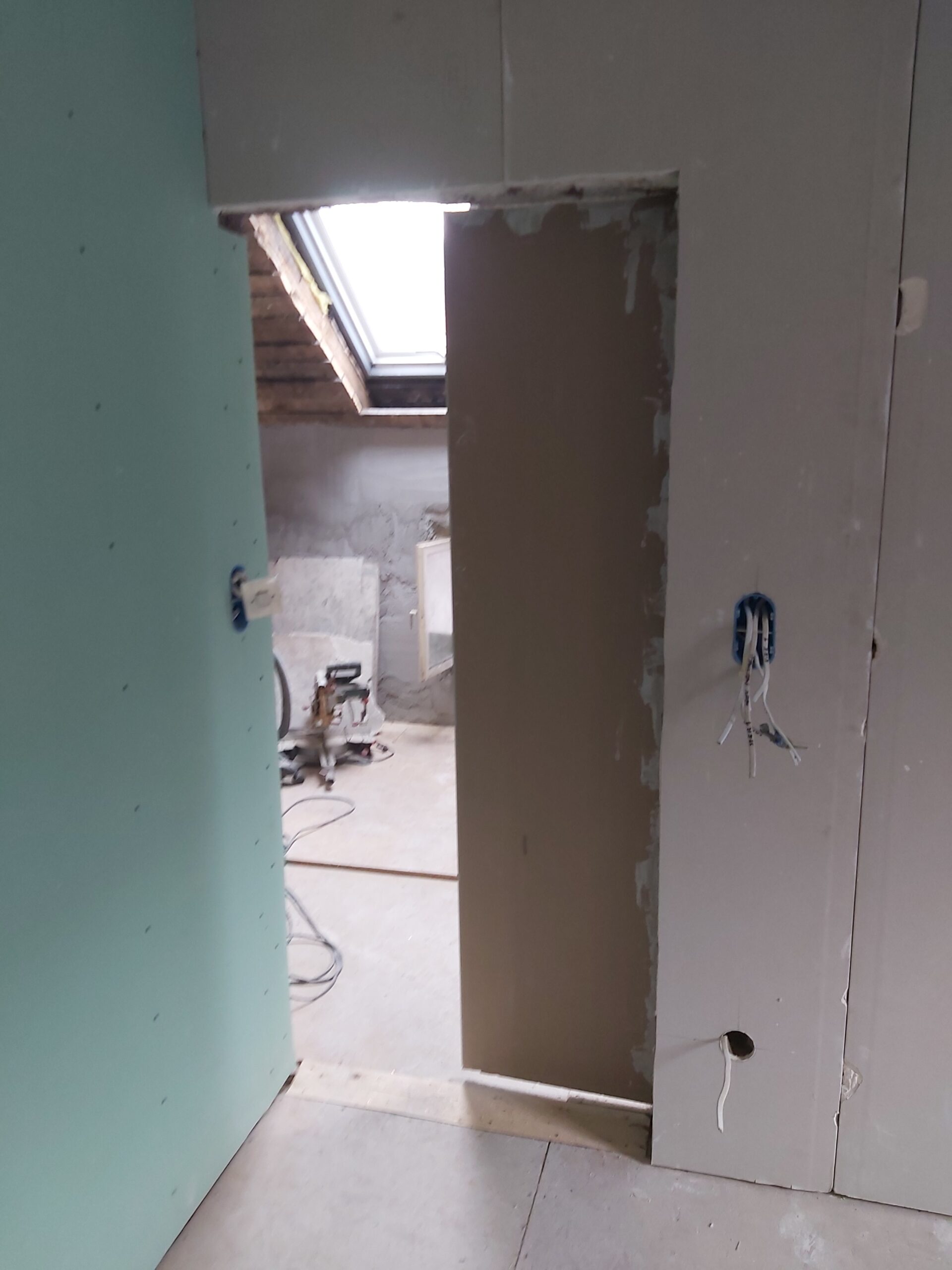 Otwór drzwiowy w trakcie remontu poddasza – ściany z płyt gipsowo-kartonowych, widoczne puszki elektryczne, sufit skośny z oknem dachowym oraz narzędzia budowlane w sąsiednim pomieszczeniu.
