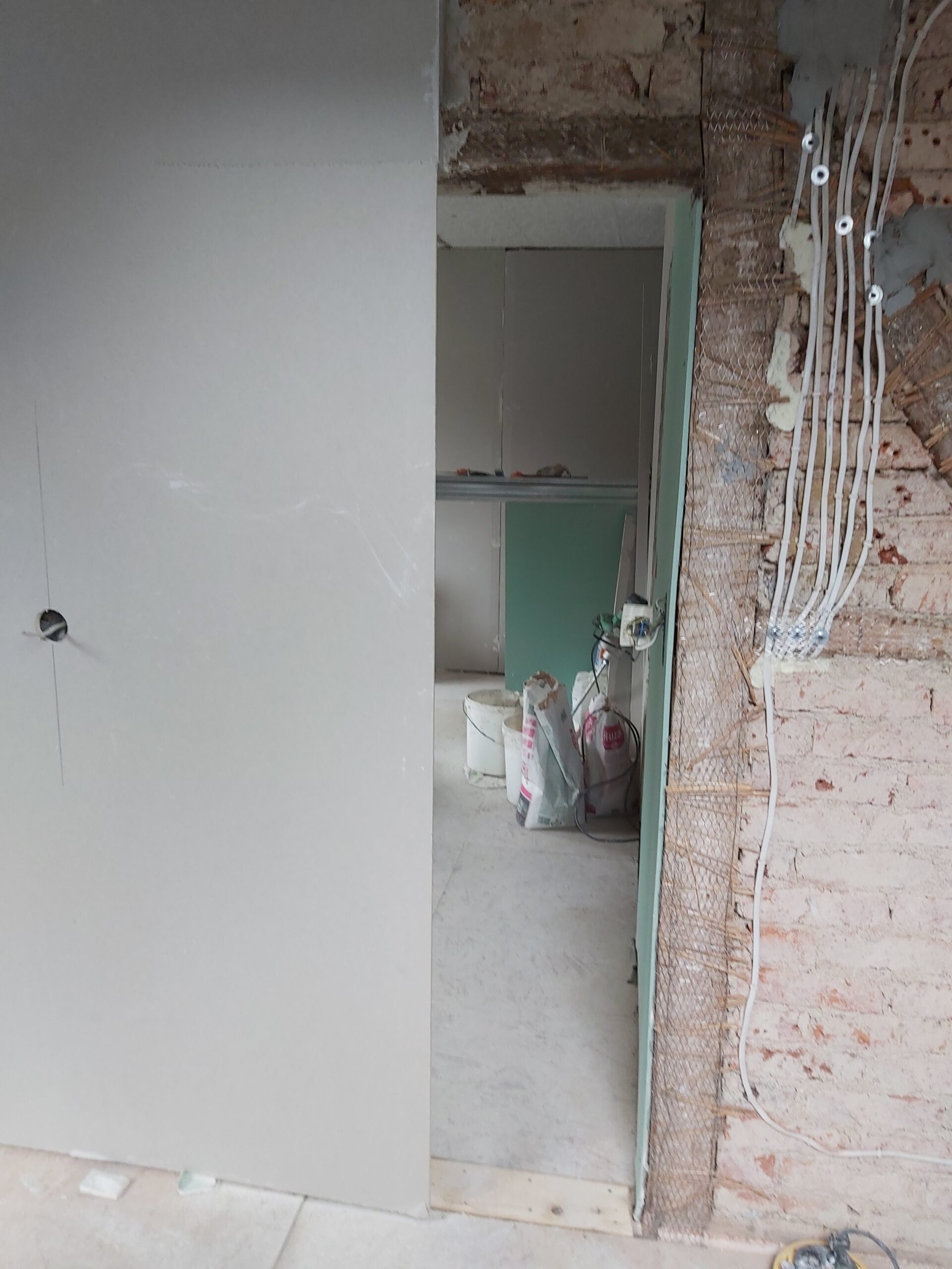 Otwór drzwiowy w trakcie remontu – ściana z płyt gipsowo-kartonowych, odsłonięta cegła z instalacją elektryczną, widoczne przewody oraz materiały budowlane w pomieszczeniu za ścianą.