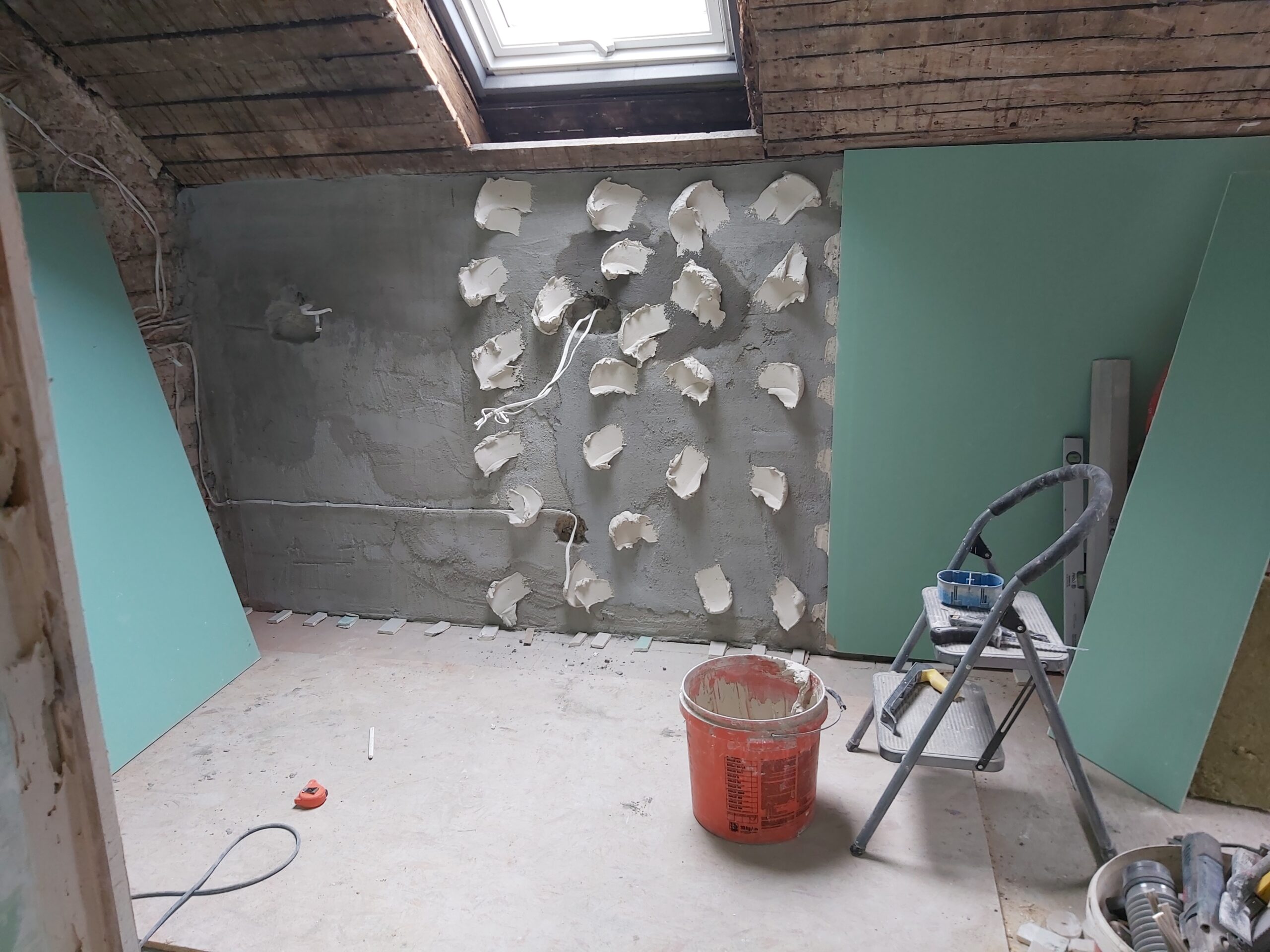 Ściana poddasza w trakcie remontu z nałożonymi punktowo plackami zaprawy klejowej, widocznym oknem dachowym, instalacją elektryczną, wiadrem z zaprawą oraz przygotowanymi płytami gipsowo-kartonowymi przed montażem.