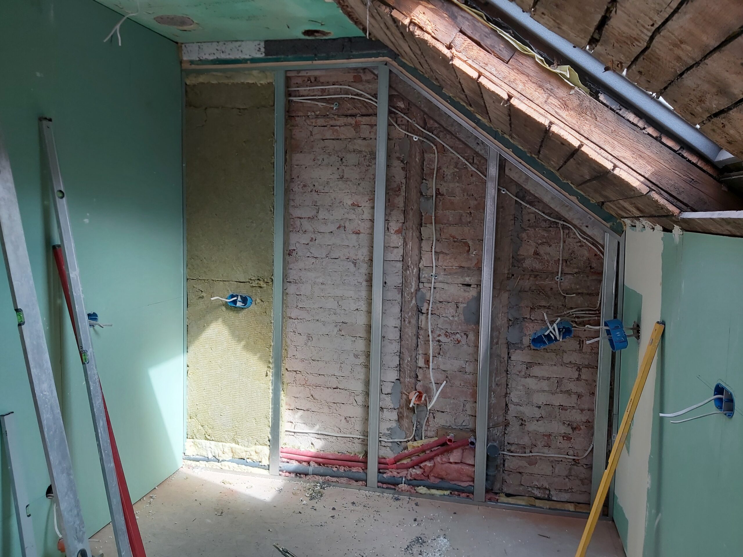 Ściana poddasza podczas remontu z widoczną stalową konstrukcją, odkrytą cegłą, prowadzeniem instalacji elektrycznej i hydraulicznej oraz częściowym ociepleniem przed zabudową płytami gipsowo-kartonowymi.