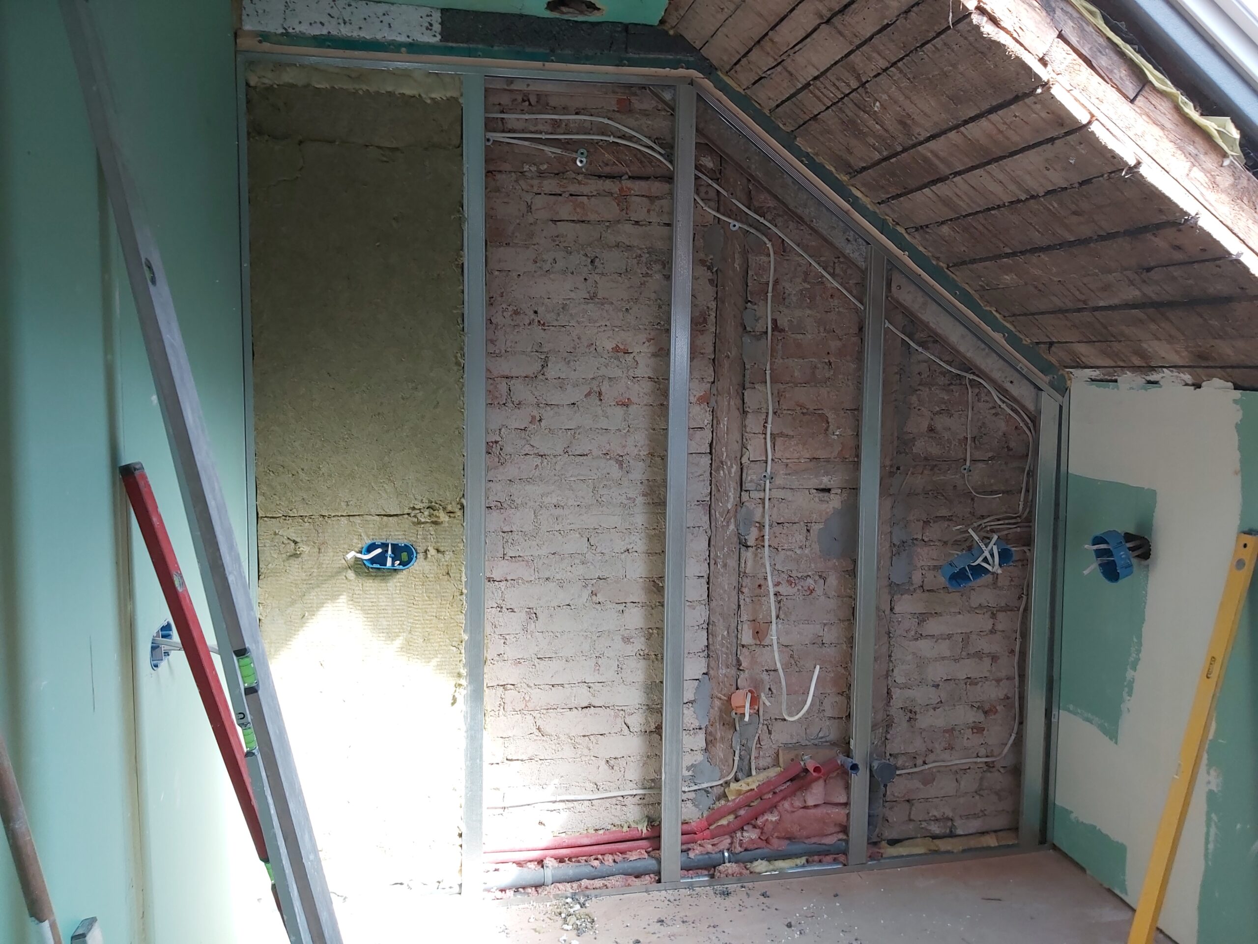 Ściana poddasza w trakcie remontu z widoczną konstrukcją stalową, odkrytą cegłą oraz poprowadzoną instalacją elektryczną i hydrauliczną, przygotowana do ocieplenia i zabudowy płytami gipsowo-kartonowymi.