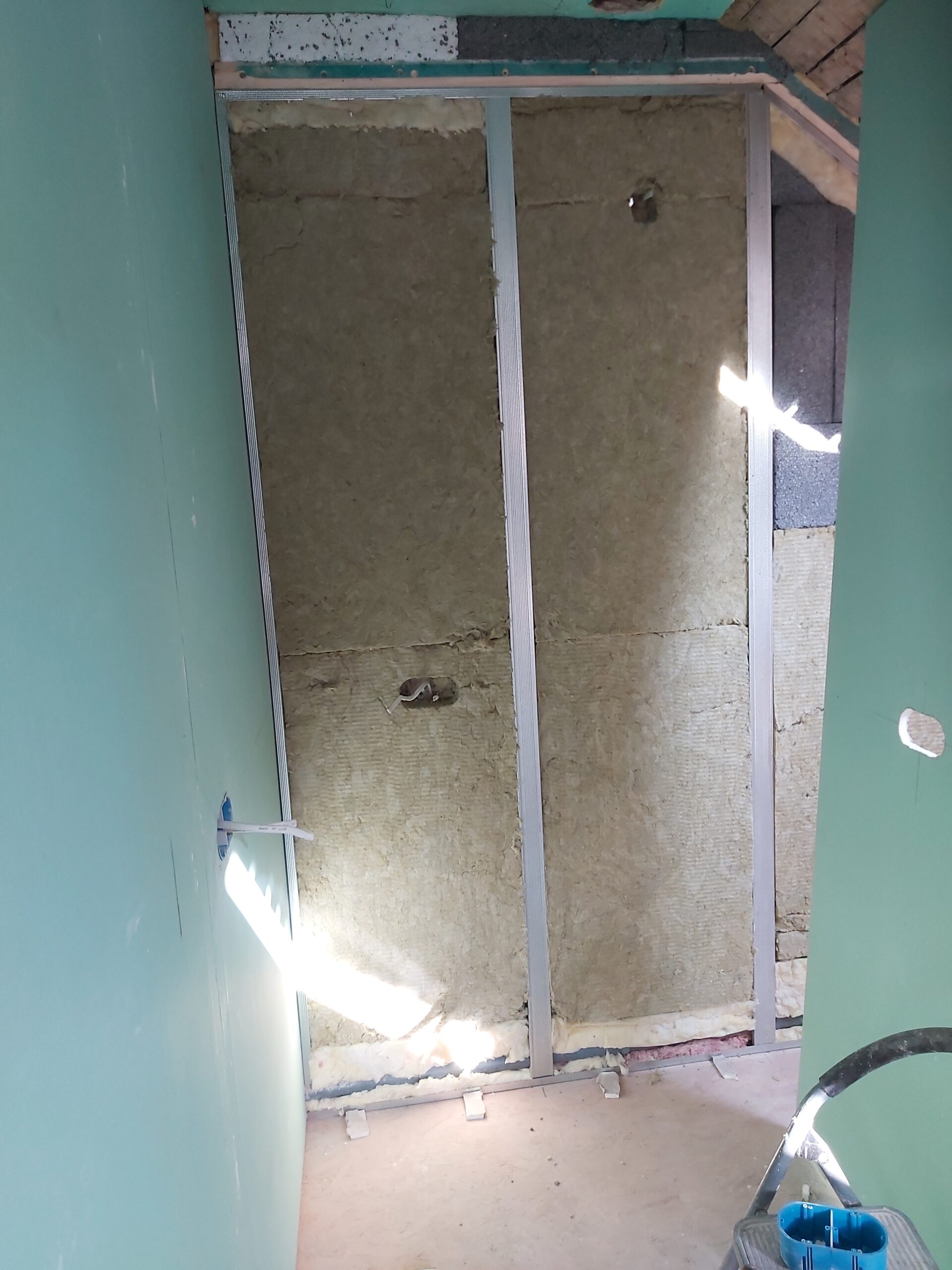 Ściana w trakcie prac wykończeniowych z widoczną konstrukcją stalową i wypełnieniem z wełny mineralnej, przygotowana do zabudowy płytami gipsowo-kartonowymi, z wyprowadzonymi punktami instalacyjnymi.