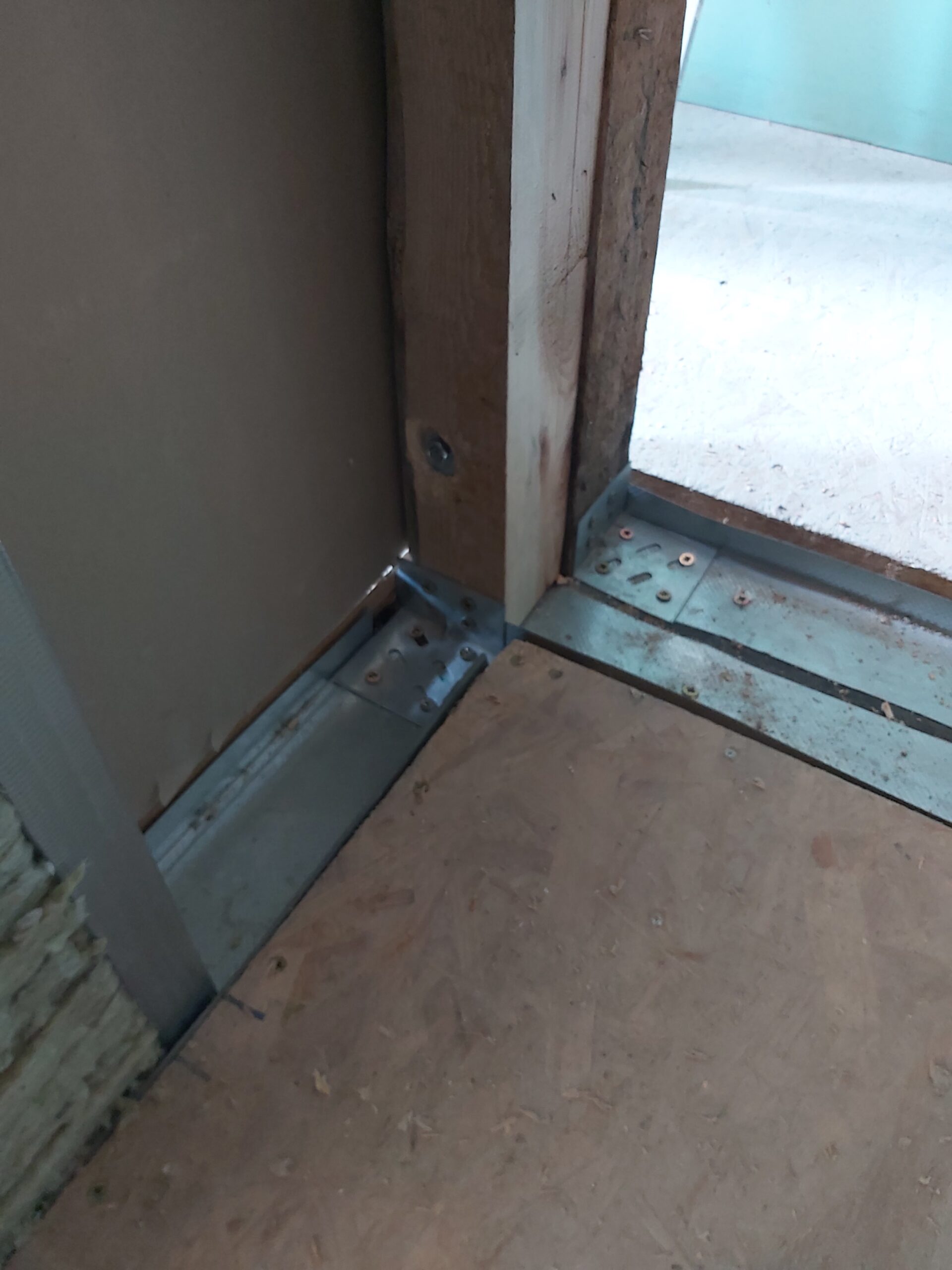 Zbliżenie na dolne połączenie profili metalowych i drewnianego słupka w konstrukcji ściany działowej, widoczne mocowania do podłogi oraz fragment poszycia podczas prac montażowych.